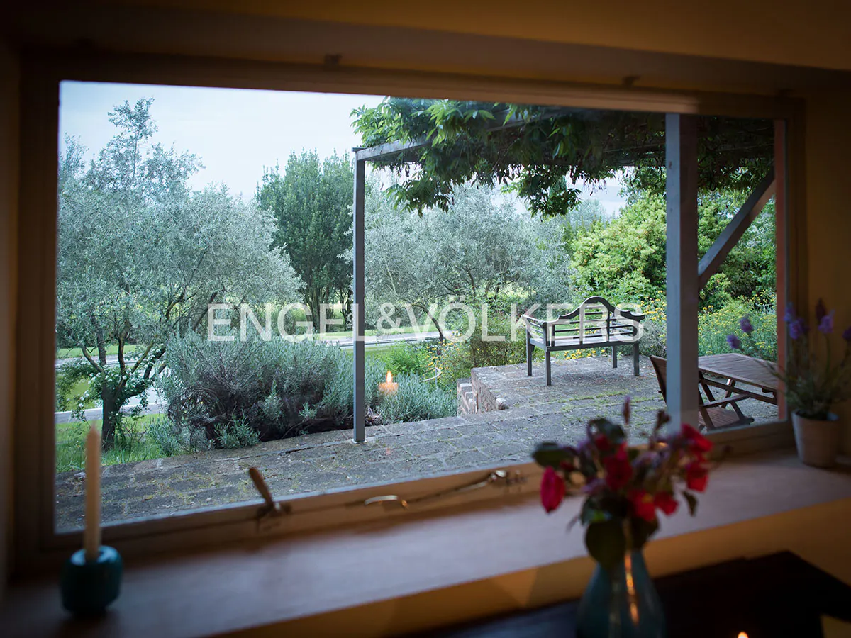 View from a window of a garden with trees, bench, and flowers. A vase of flowers sits on the windowsill.