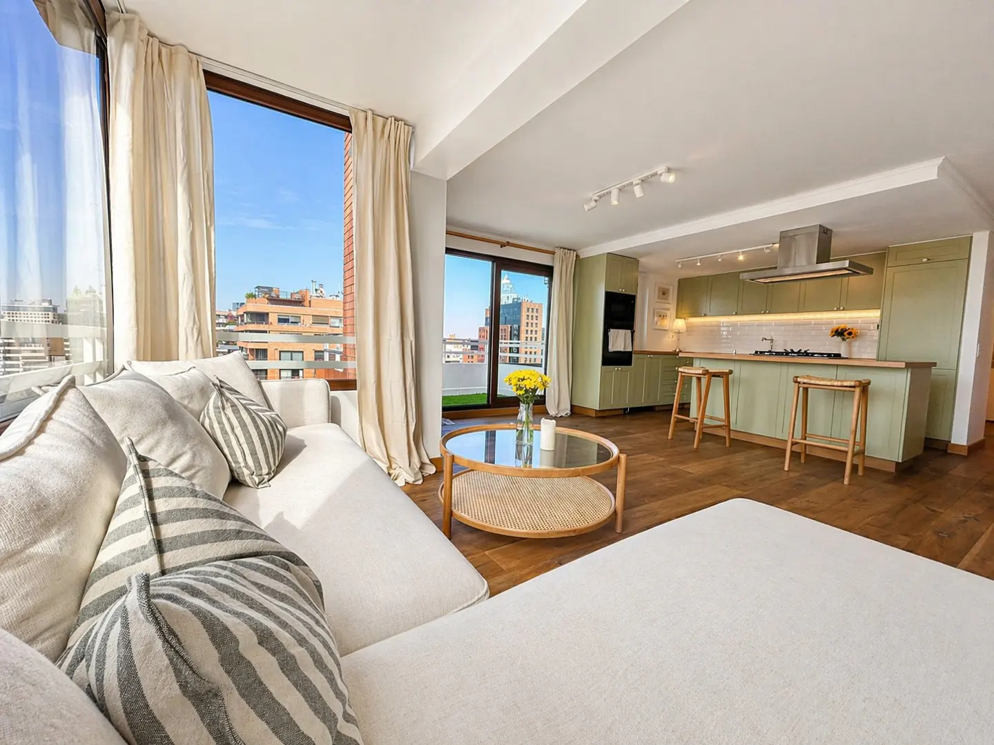 Bright, modern apartment interior with a white sofa, round coffee table, and a green kitchen in the background. Large windows offer a city view.