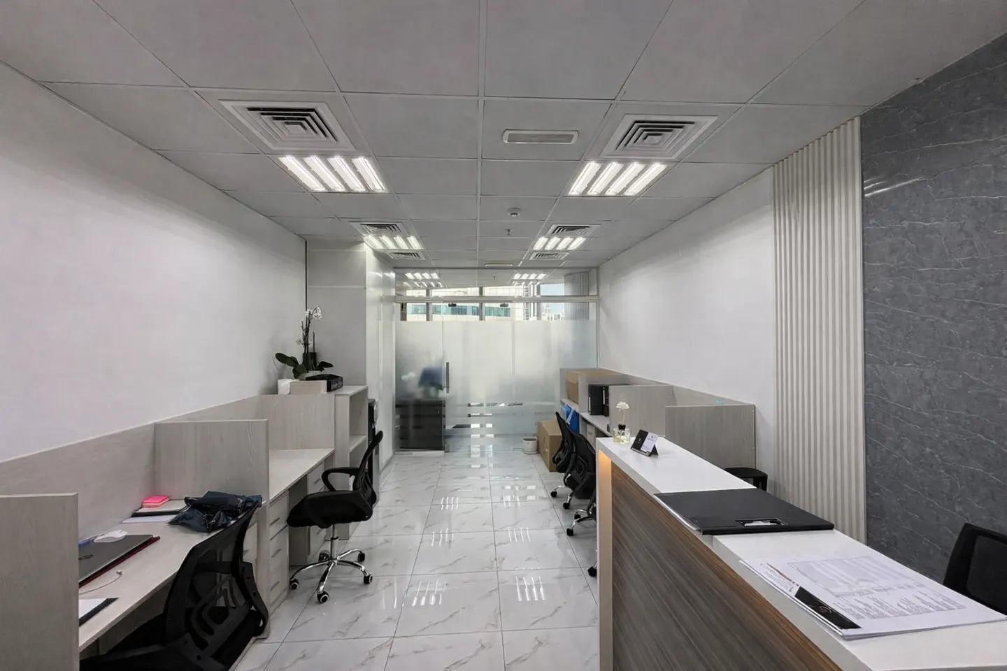 Modern office space with white marble floors, rows of workstations, black chairs, and a reception desk.