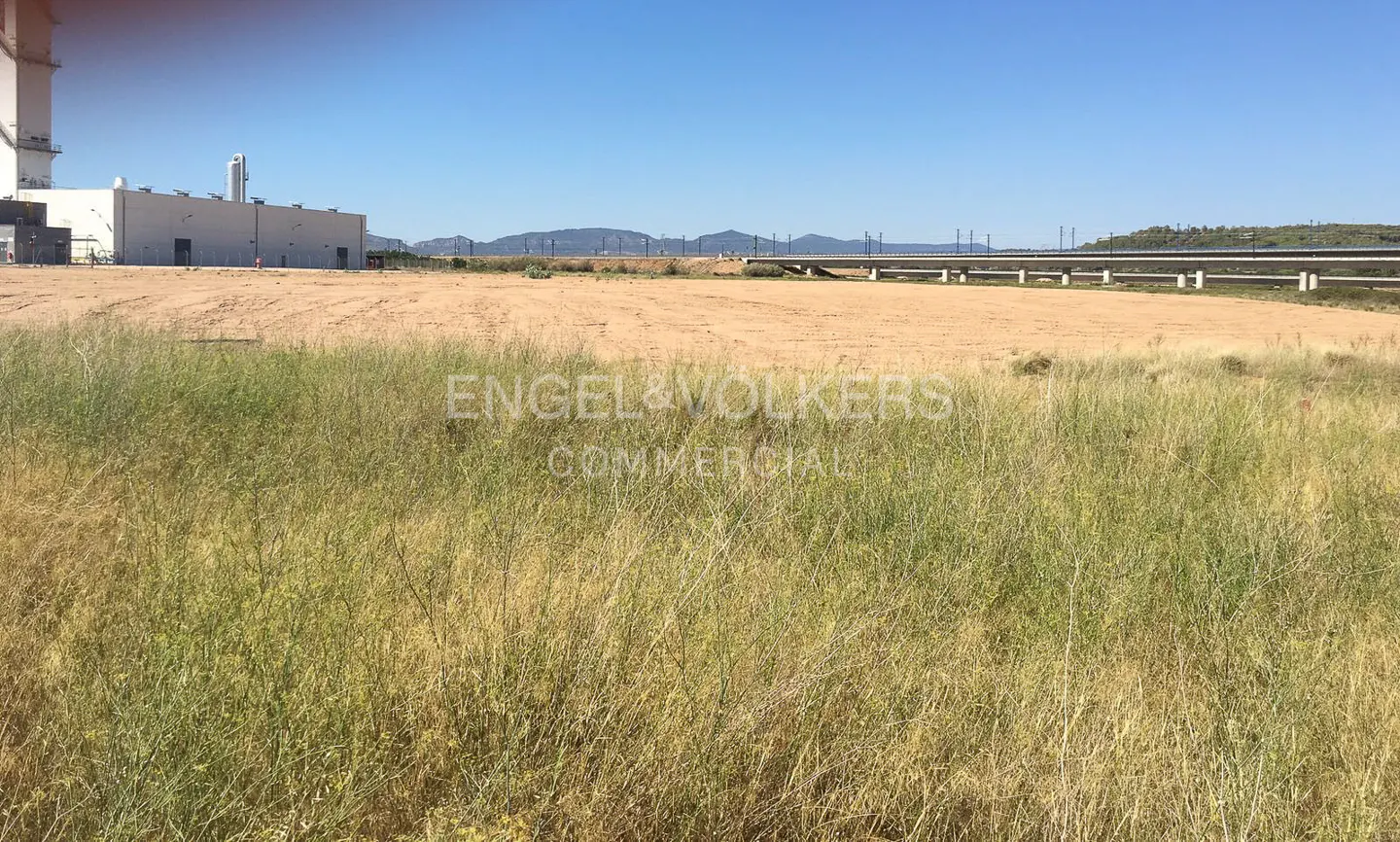 Large vacant lot with dry grass, a white industrial building, and a distant bridge under a clear blue sky.