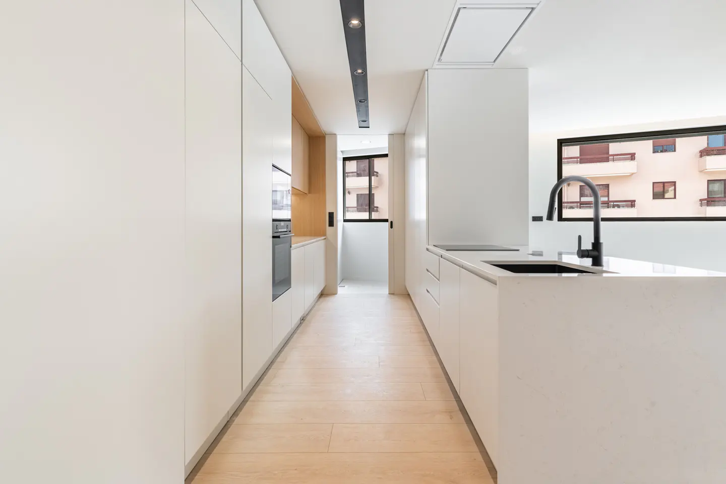 Modern galley kitchen with white cabinets, light wood floors, a black faucet, and a view of an apartment building outside.