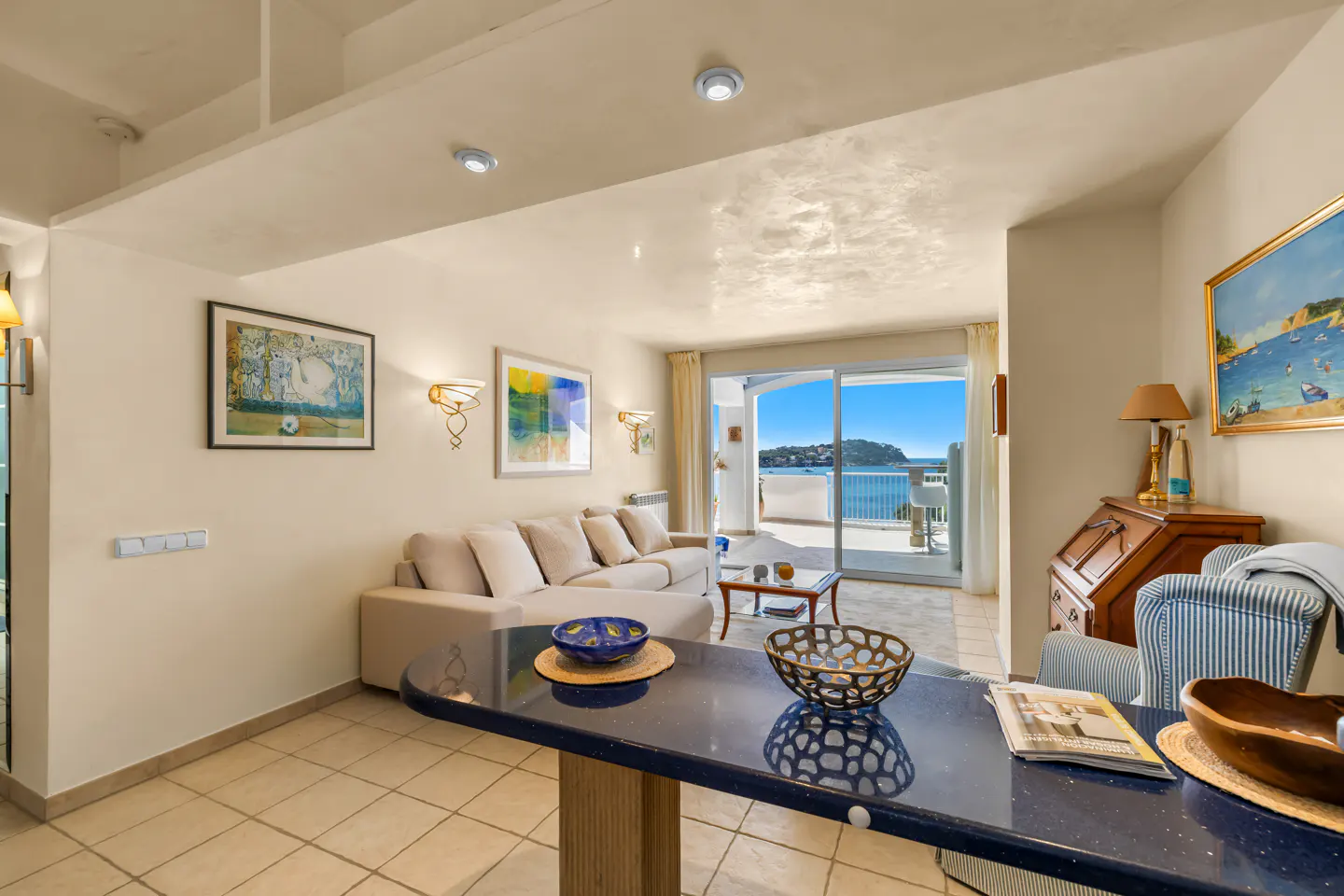 Bright living room with beige sofa, blue counter, and ocean view through sliding glass doors. Artwork and lamps decorate the walls.