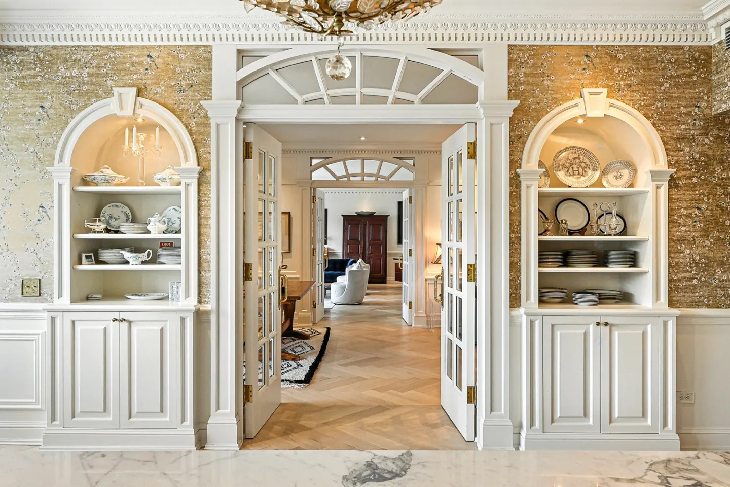 Elegant entryway with open French doors, flanked by white built-in shelves displaying china. Floral wallpaper and herringbone floors add charm.
