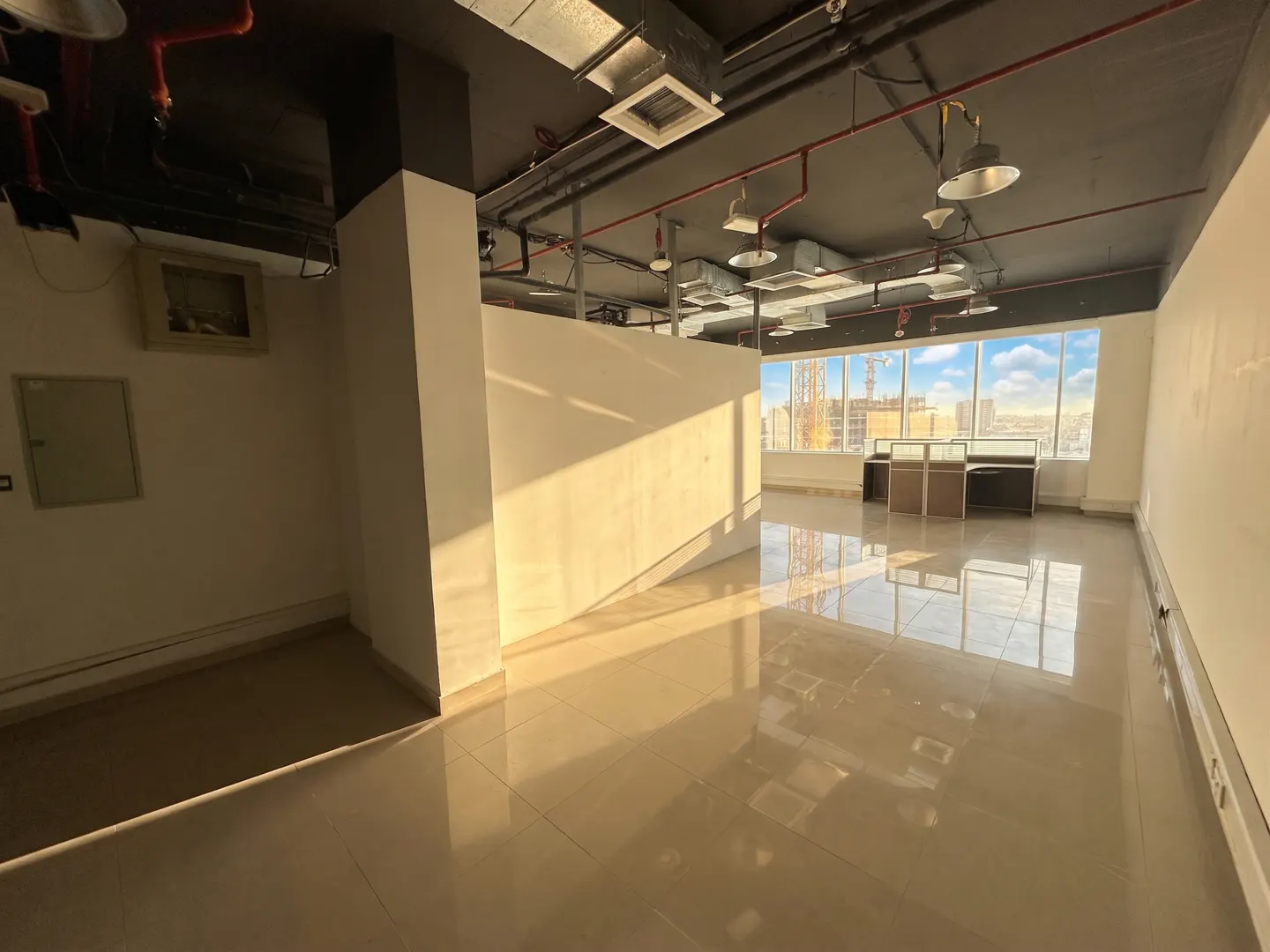 An empty office space with beige tile floors, white walls, and a black ceiling with exposed pipes and lights. Large windows offer a city view.