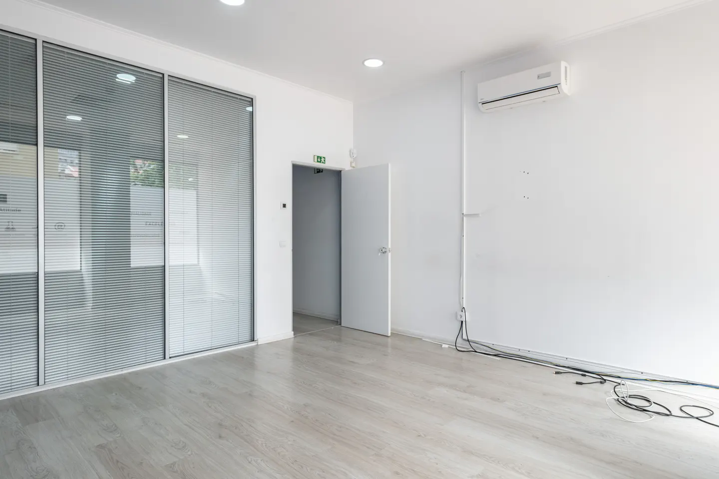 Bright, empty office space with light wood floors, white walls, and large glass windows with blinds. A white door is slightly ajar.