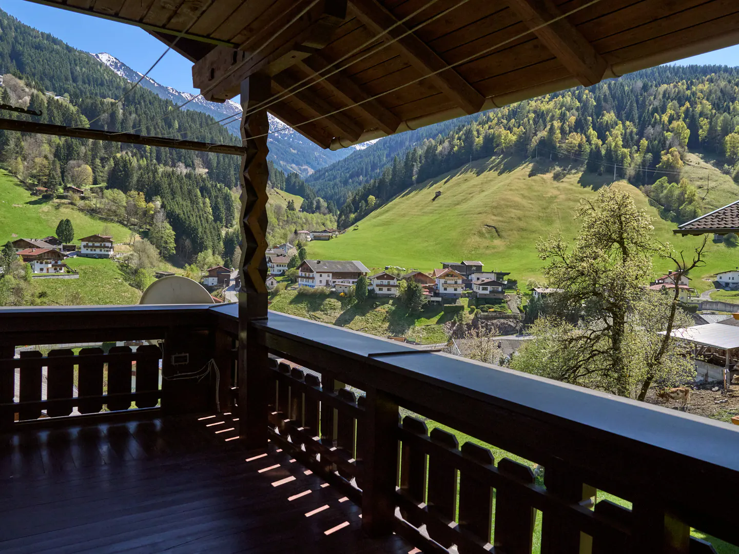 View from a wooden balcony overlooking a green valley with houses and mountains in the background.