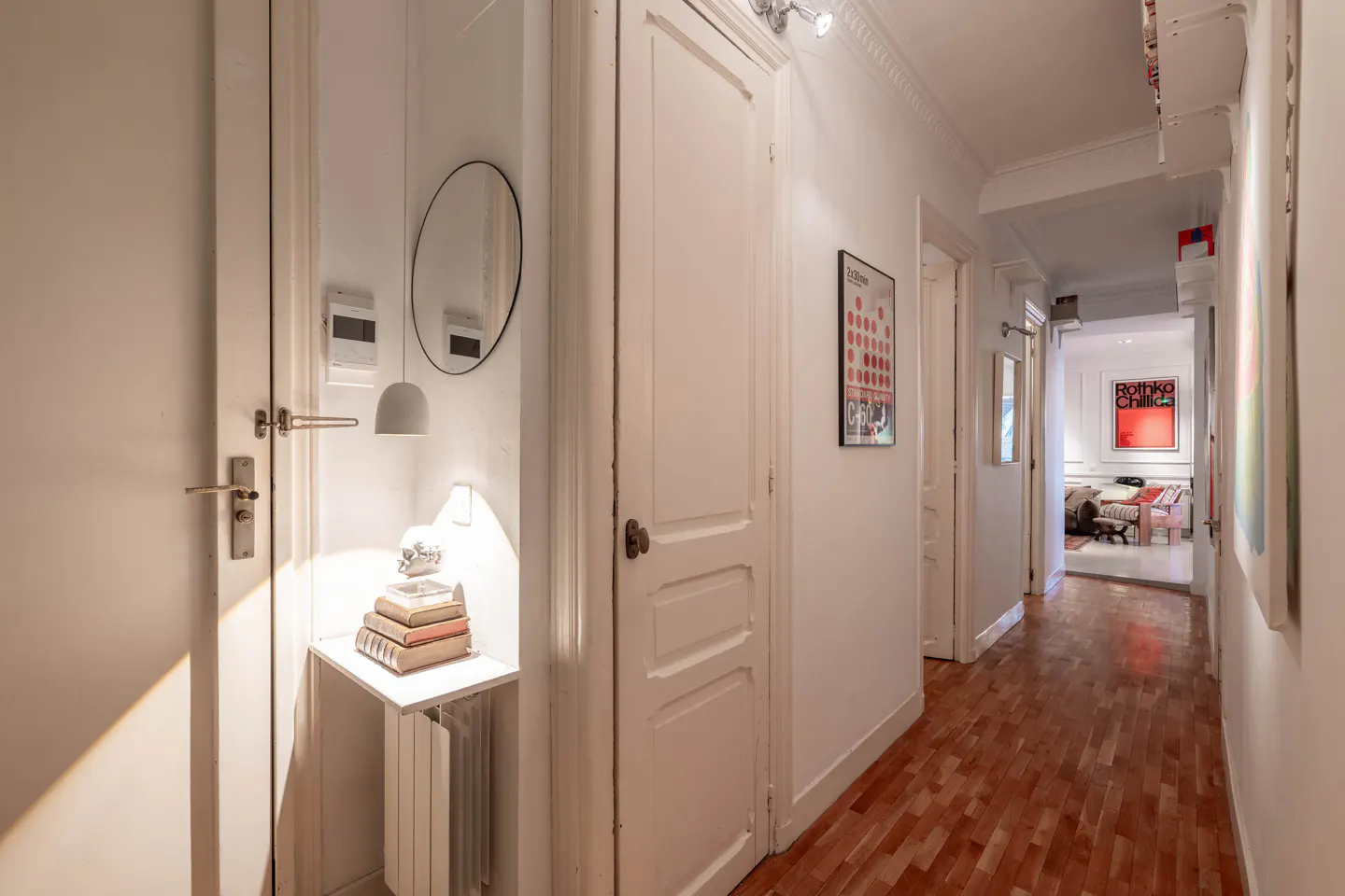 Hallway with white walls, wood floors, and doors. A small shelf holds books, a lamp, and a round mirror. Artwork hangs on the walls.