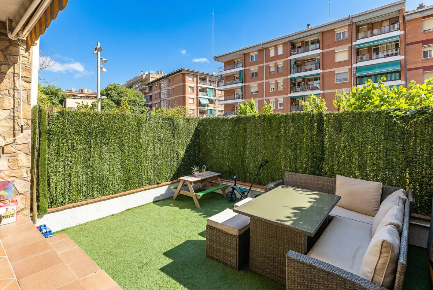 A cozy outdoor patio with green artificial turf, a wicker sofa, and a picnic table, surrounded by a tall green hedge.
