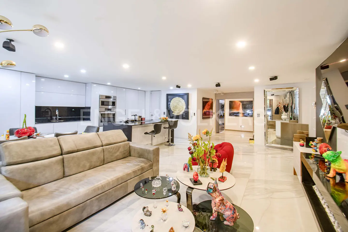 Open-concept living space with a beige sofa, glass tables, and a modern white kitchen with a black island and bar stools. Artwork adorns the walls.