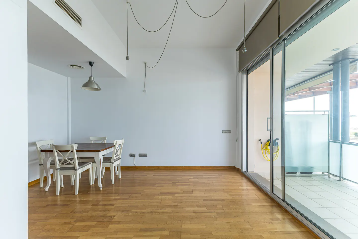 Minimalist dining area with a wooden table, four white chairs, and a pendant light, next to a large glass balcony door.