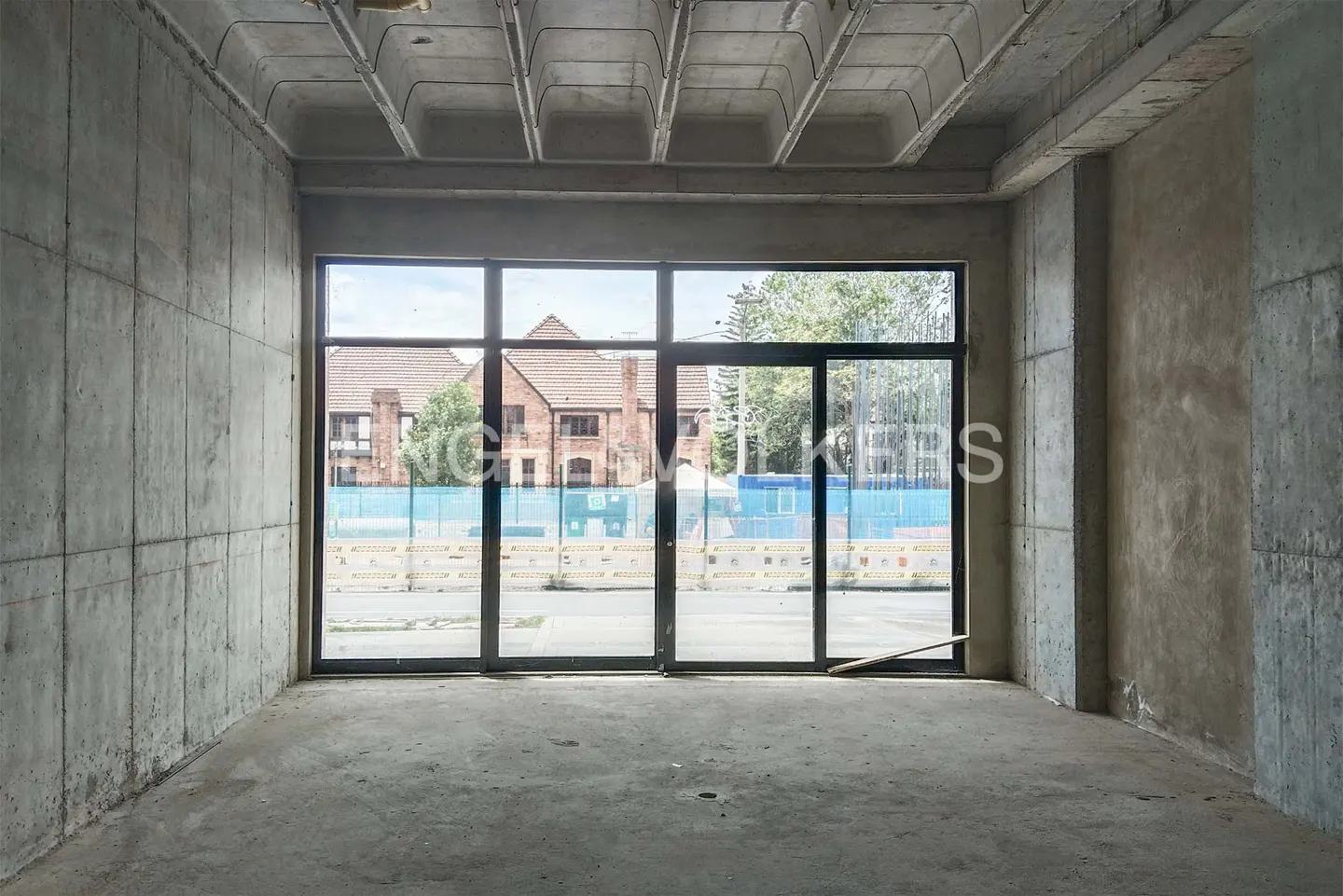 Unfinished room with concrete walls, floor, and ceiling. Large window shows a brick building and construction fencing outside.