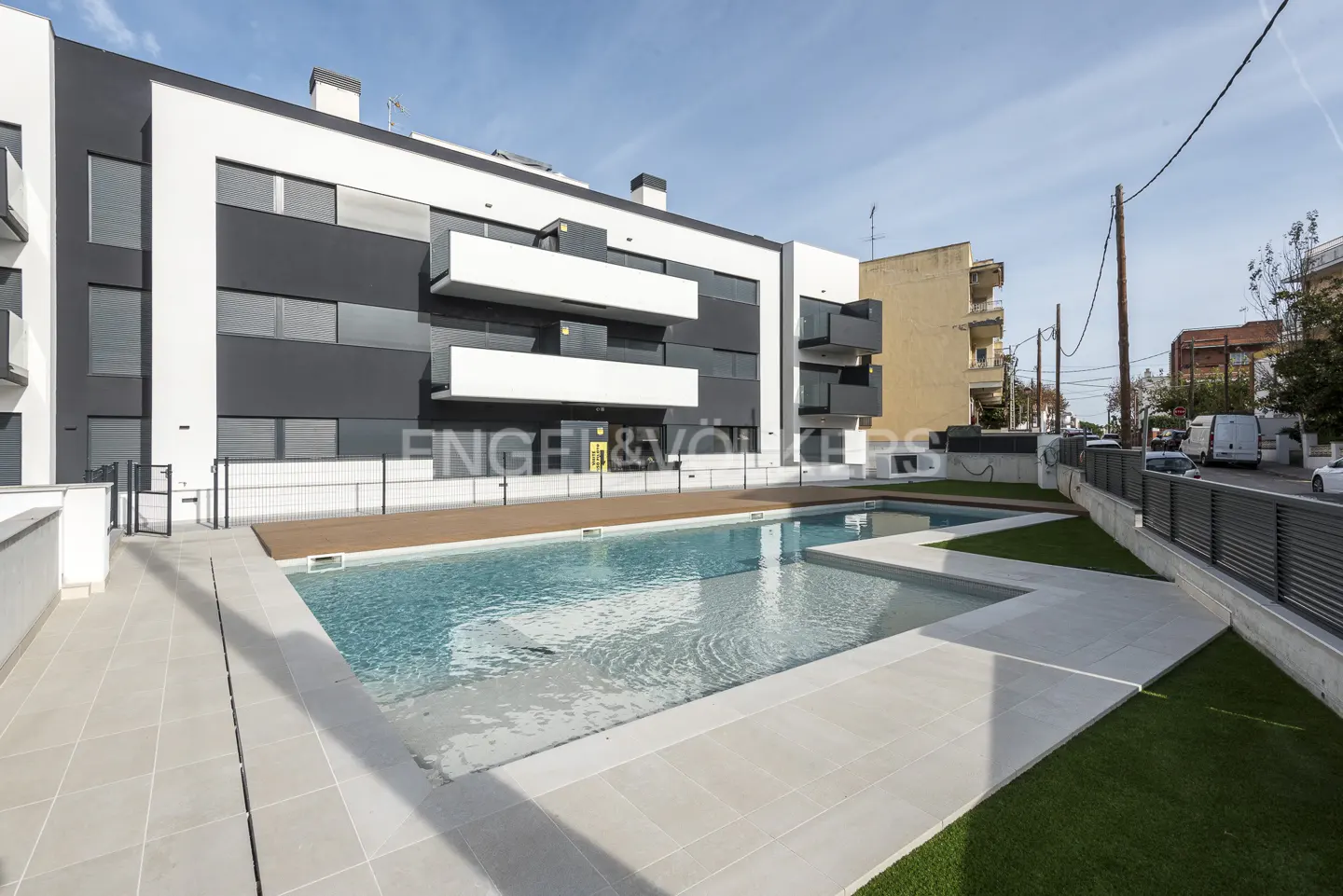 Modern apartment building with a pool. The building is white and gray with balconies. The pool is surrounded by tile and artificial grass.