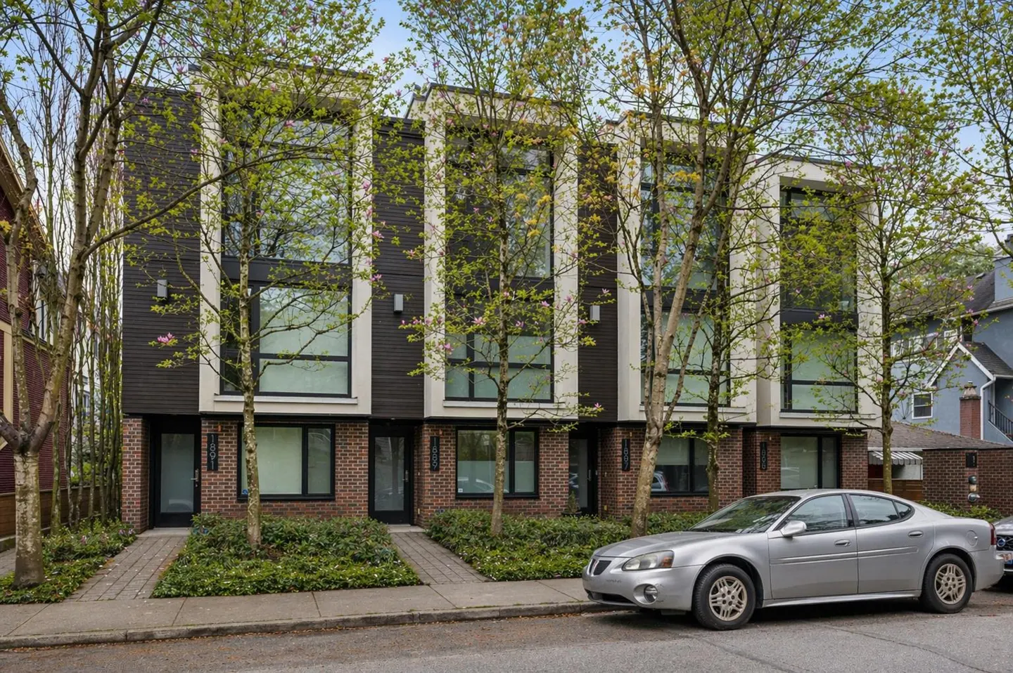 Three modern townhouses with dark brown siding, brick bases, and large windows, framed by trees. A silver car is parked on the street.