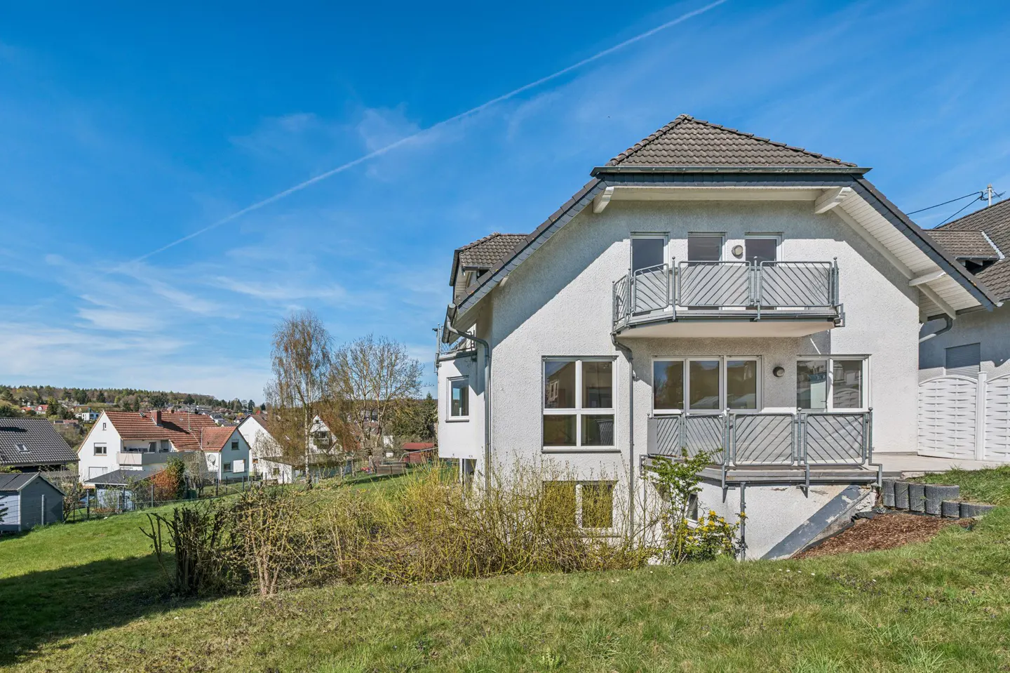 Exterior view of a white house with a brown roof and two balconies, set on a grassy hill under a blue sky.