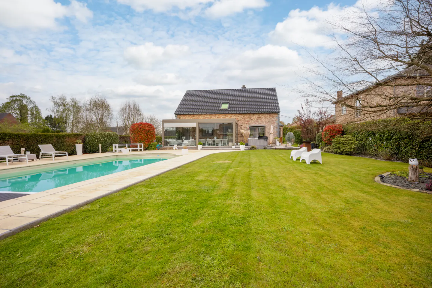 A backyard with a pool, green lawn, and a stone house with a glass-enclosed patio under a cloudy sky.