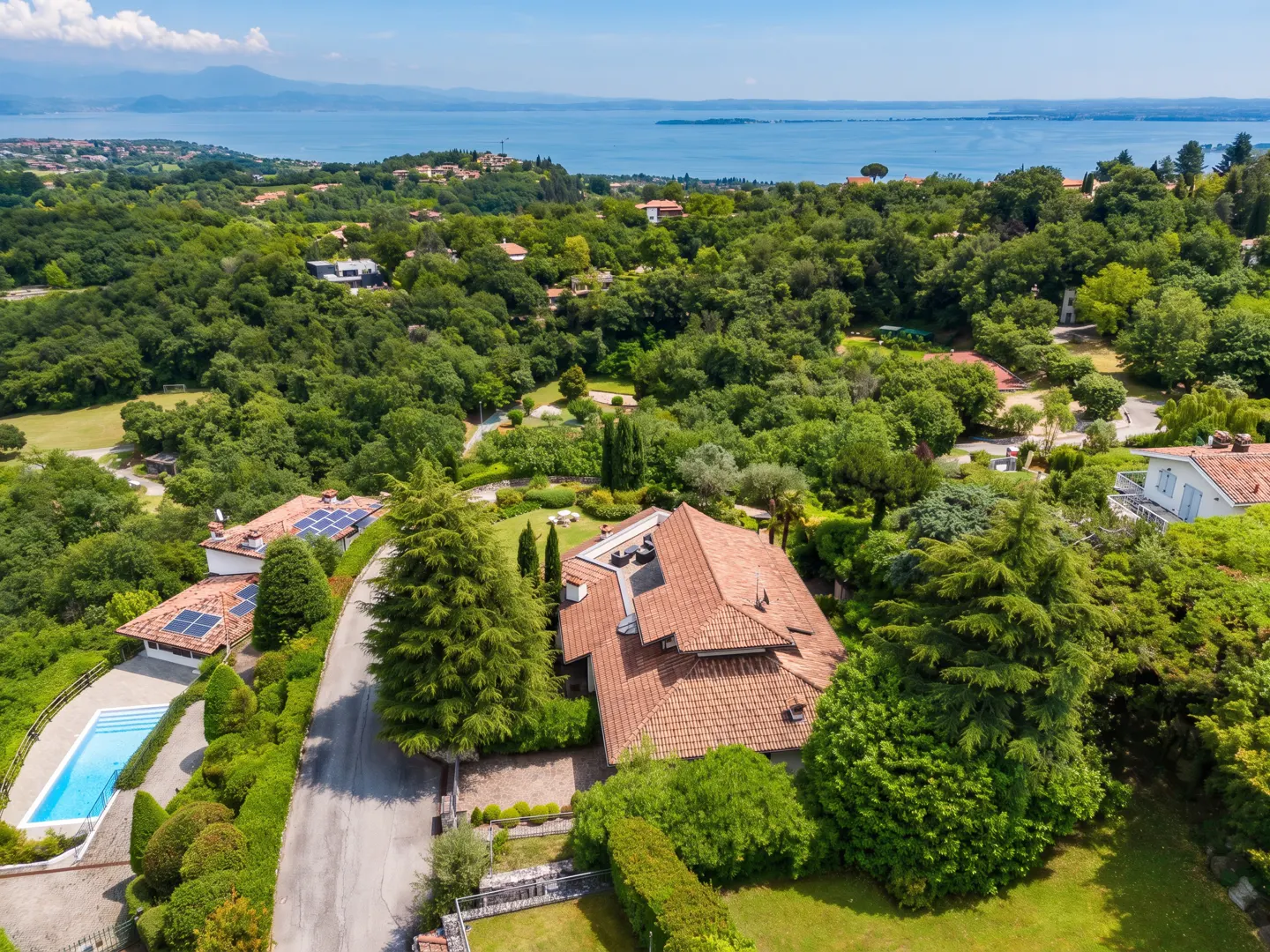 Aerial view of a terracotta-roofed house surrounded by green trees, with a blue lake and mountains in the background.