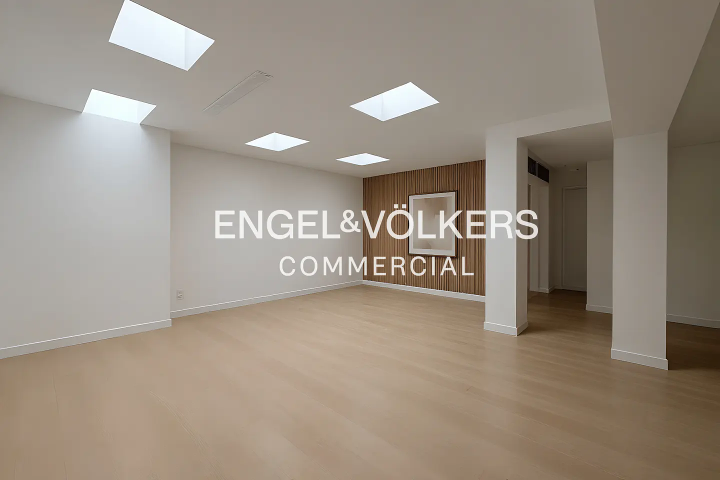 Bright, empty commercial space with light wood floors, white walls, and skylights. A wood-paneled wall features a framed picture.