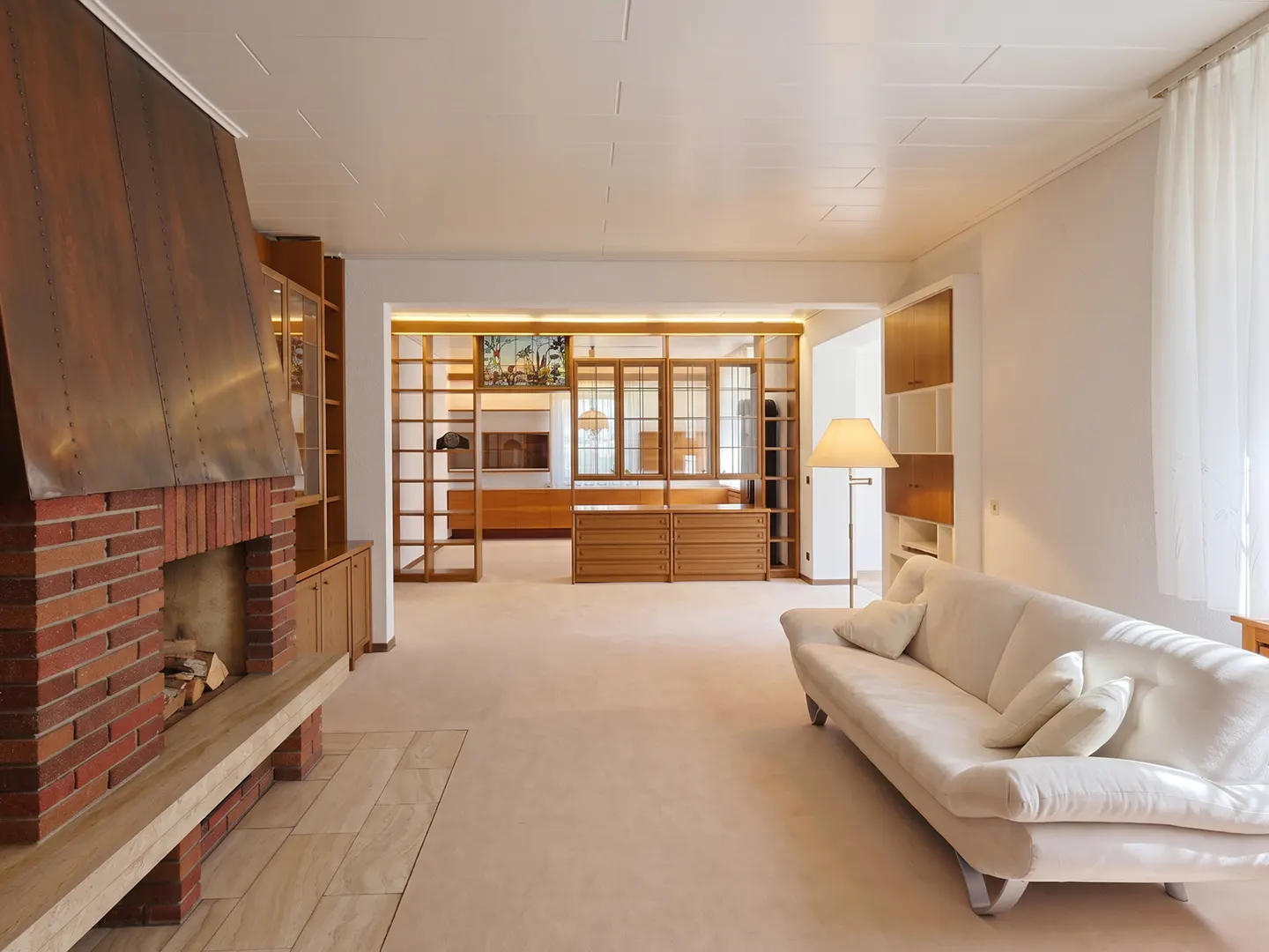 A bright living room with a brick fireplace, a white sofa, and wooden cabinets. The room is spacious and well-lit, with a neutral color scheme.