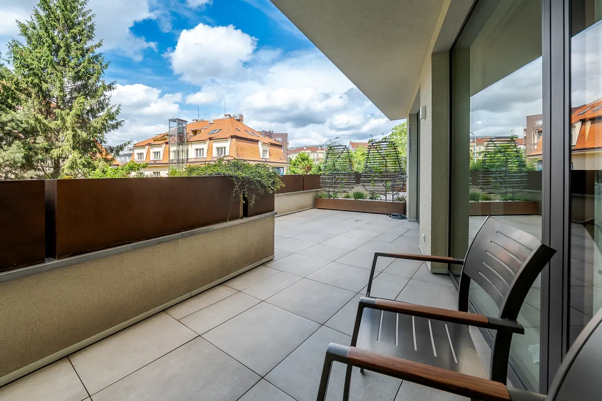 A modern balcony with gray tile flooring, metal chairs, and planters with greenery. Buildings and trees are visible in the background under a blue sky.