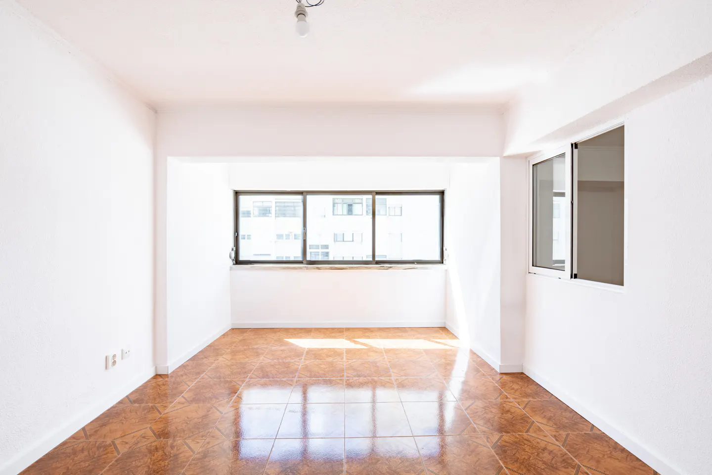 Bright, empty room with white walls and brown tile floor. Large windows let in sunlight. A lightbulb hangs from the ceiling.