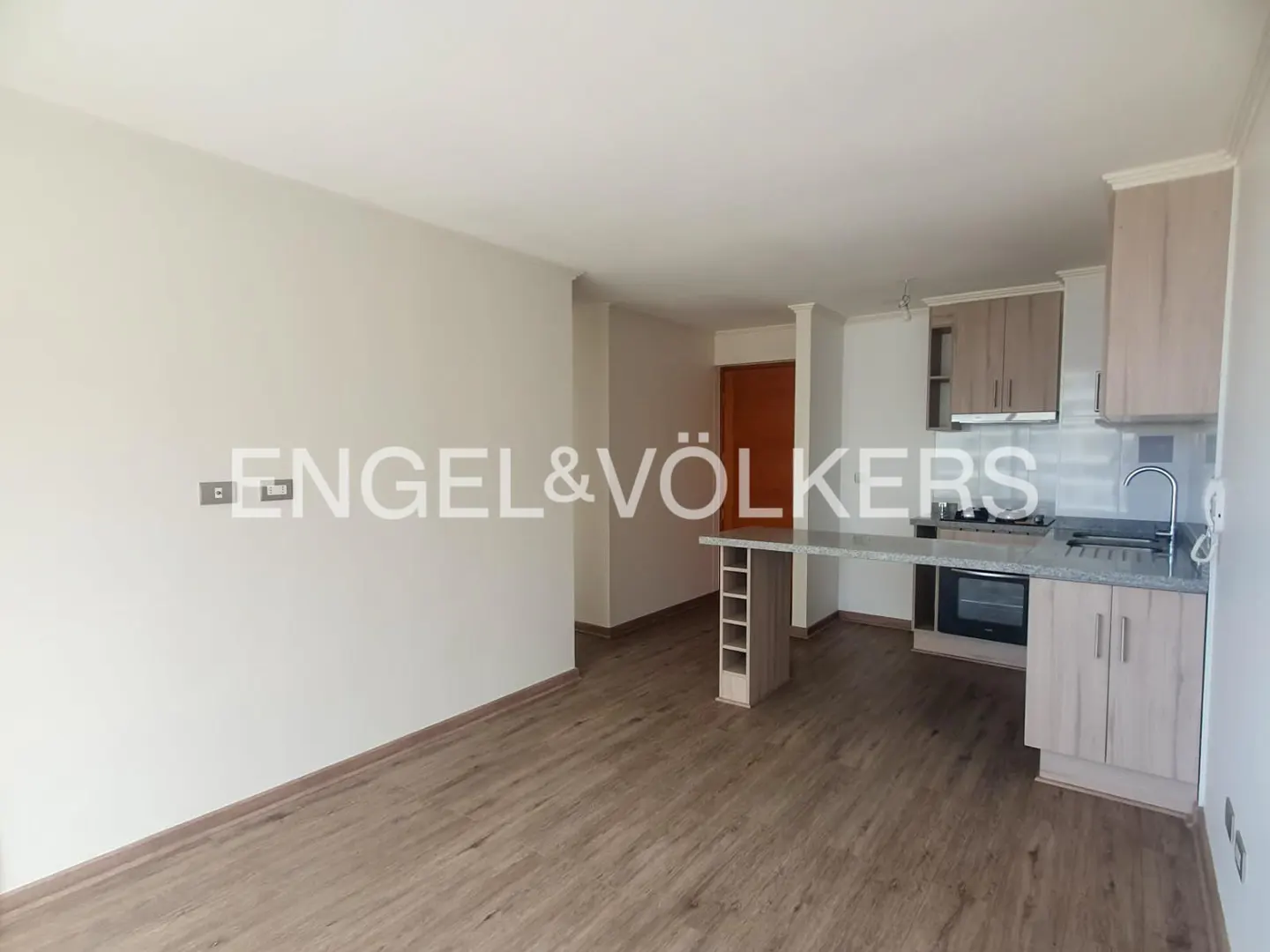 A bright, open-concept apartment with wood floors, a kitchen with light wood cabinets, and a gray countertop. The Engel & Völkers logo is visible.
