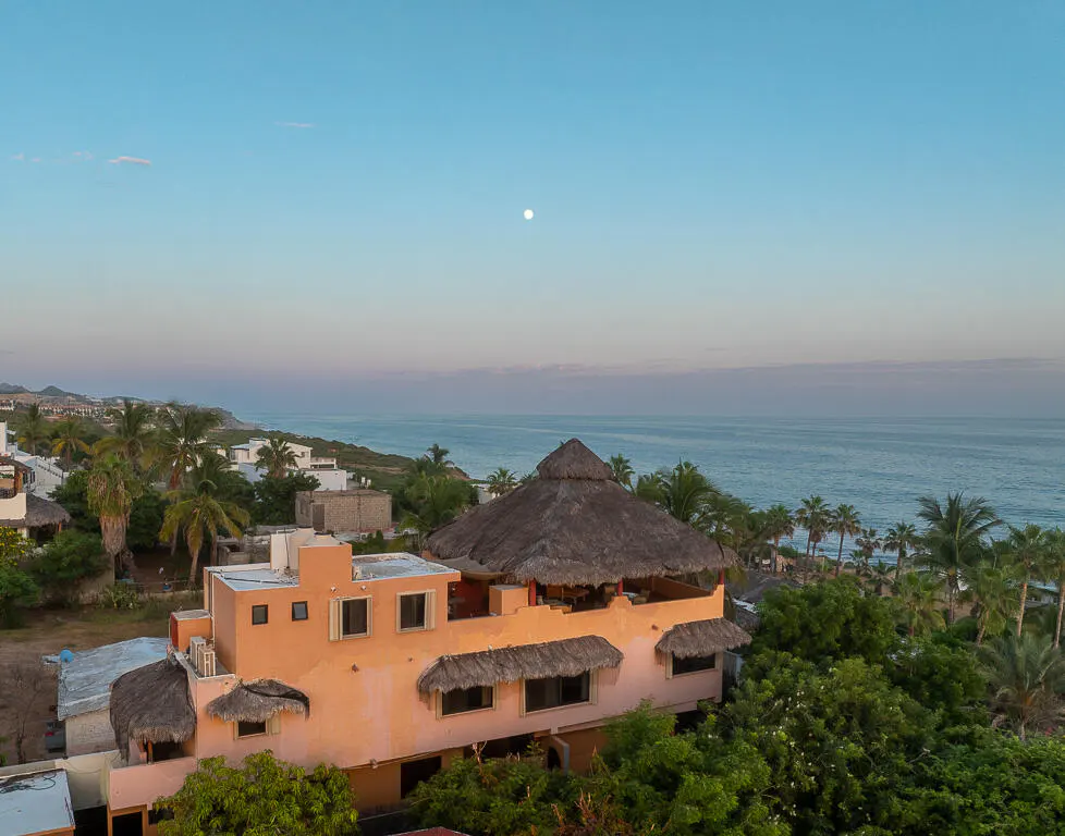 Coastal view of a peach-colored building with a thatched roof, palm trees, and the ocean under a blue sky with a visible moon.