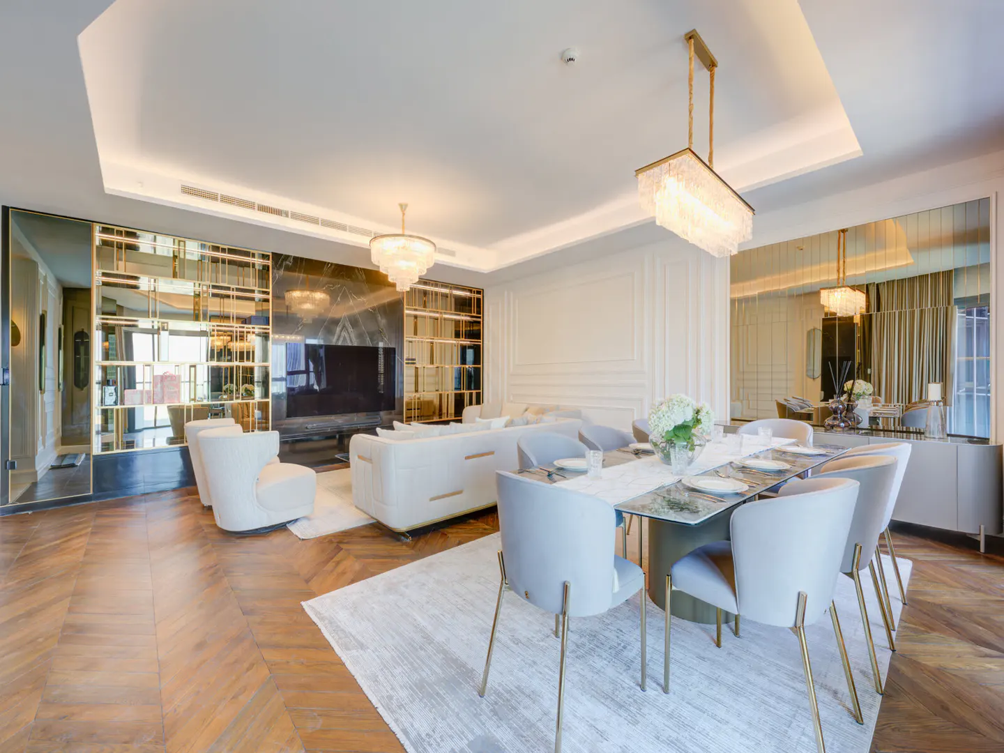 A bright, modern living and dining area with white sofas, gray chairs, and a glass-topped table. A chandelier hangs above the table.