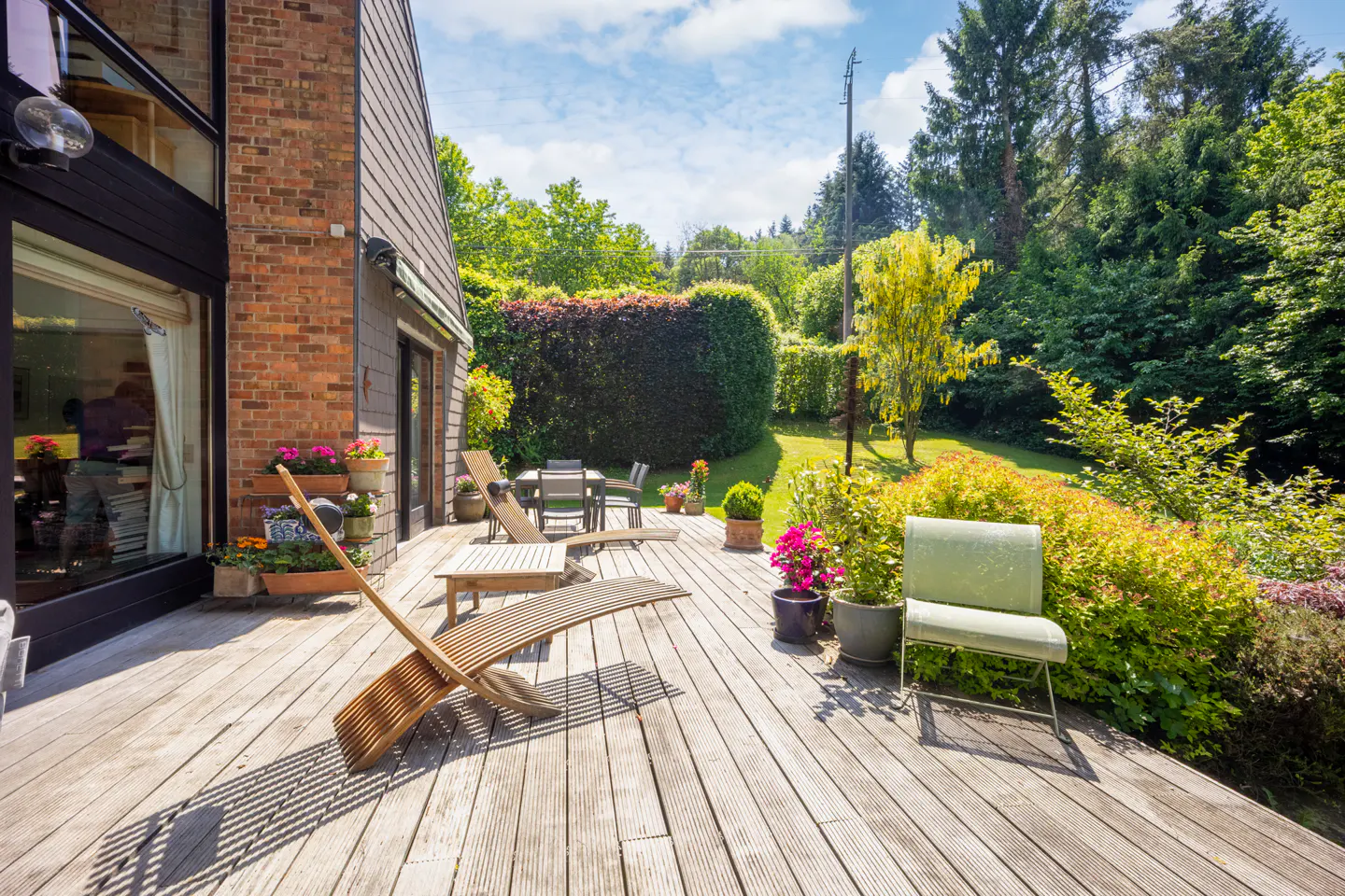 Wooden deck with lounge chairs, table, and green chair. Brick and siding house with garden and trees in the background.