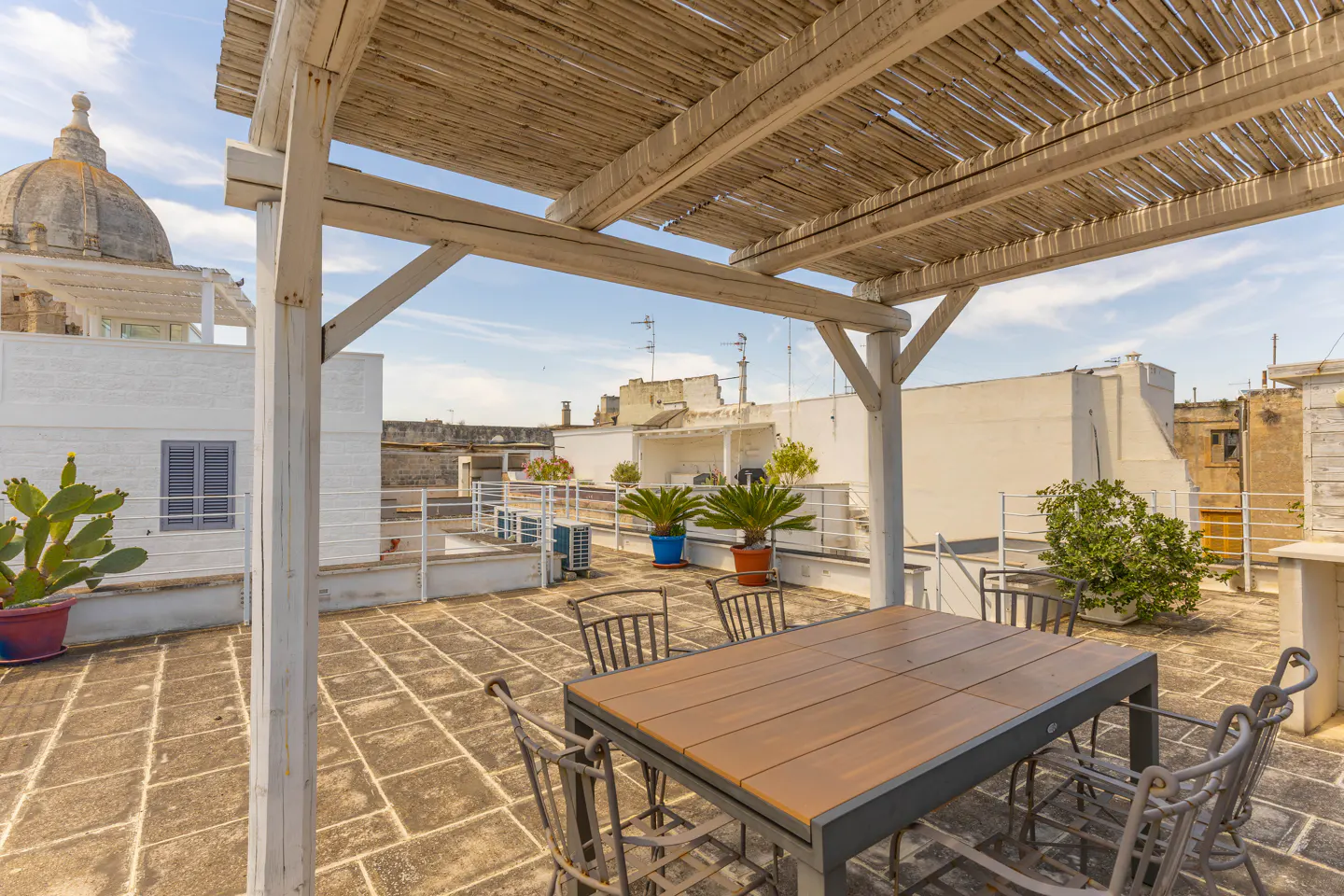 Rooftop terrace with a wooden pergola, dining table, chairs, potted plants, and a view of historic white buildings.