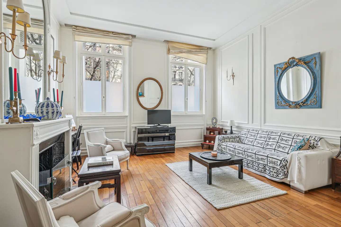 Elegant living room with hardwood floors, white walls, a patterned sofa, two armchairs, and a fireplace with a mirror.