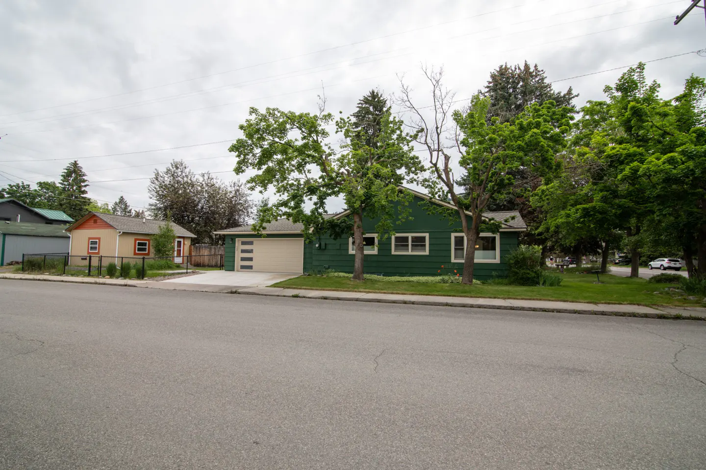A green single-story house with a tan garage next to a smaller tan house on a quiet street under a cloudy sky.