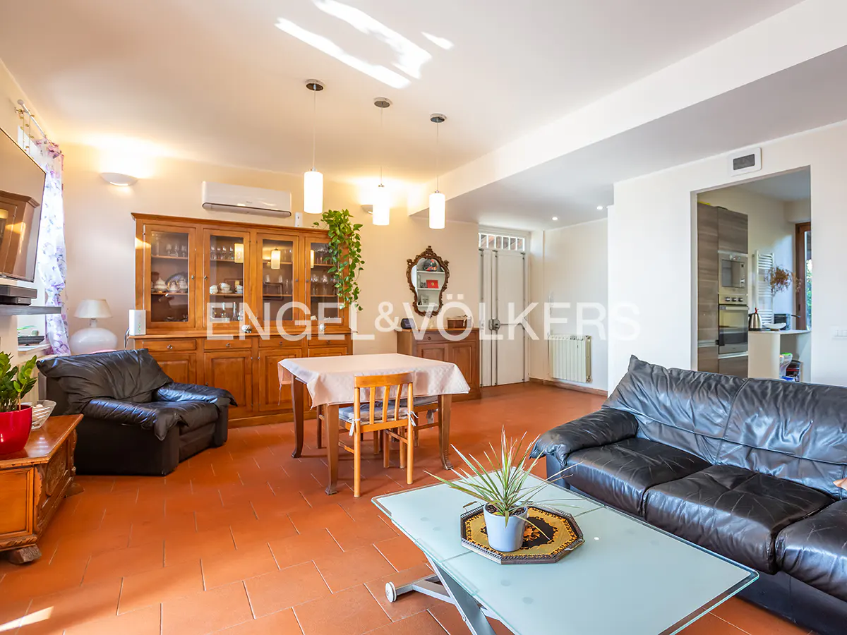 A bright living room with terracotta tile floors, a wooden dining table, and a black leather sofa.