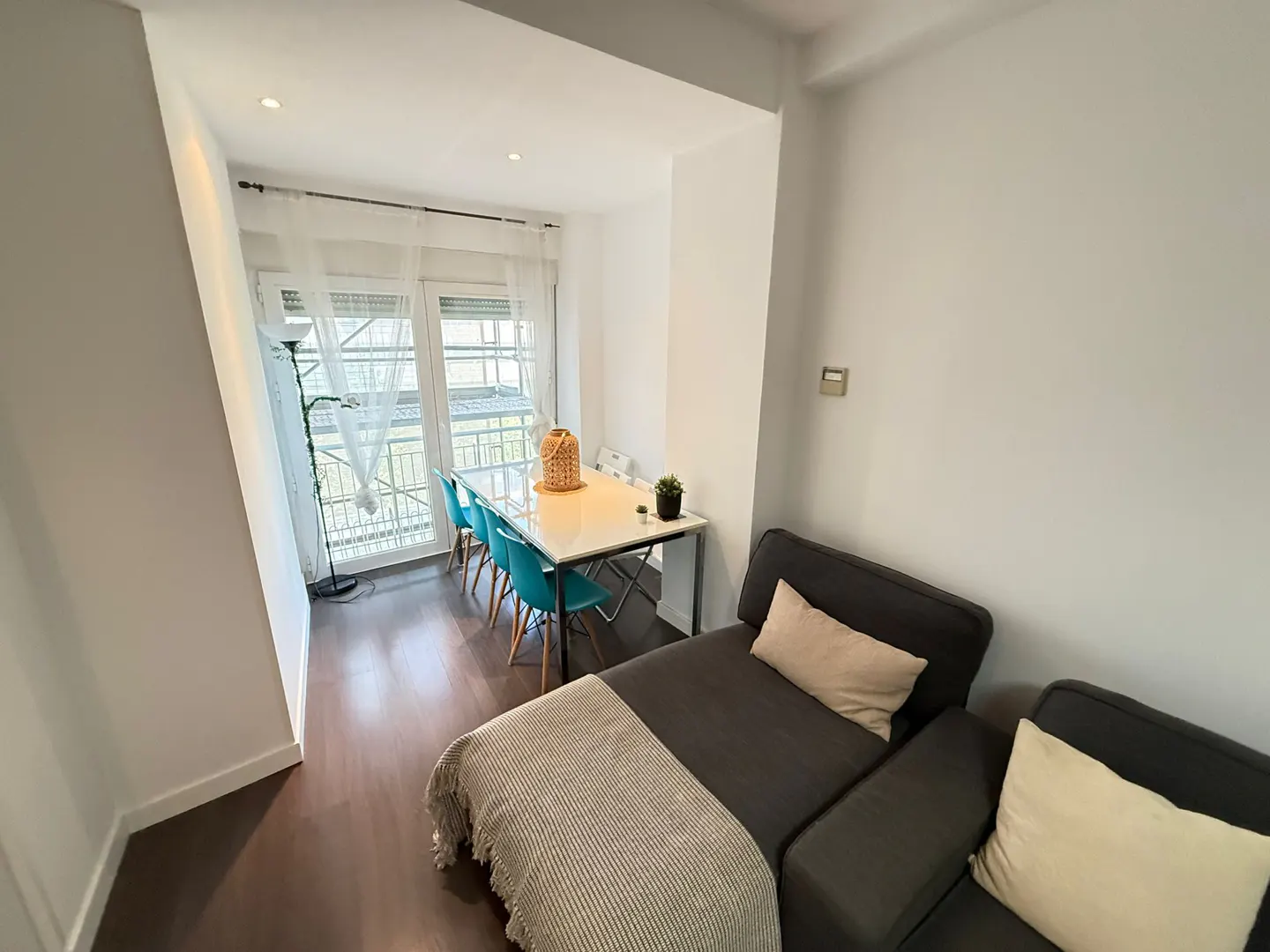 Living room with a gray sofa, a white table with blue chairs, and a window with white curtains.