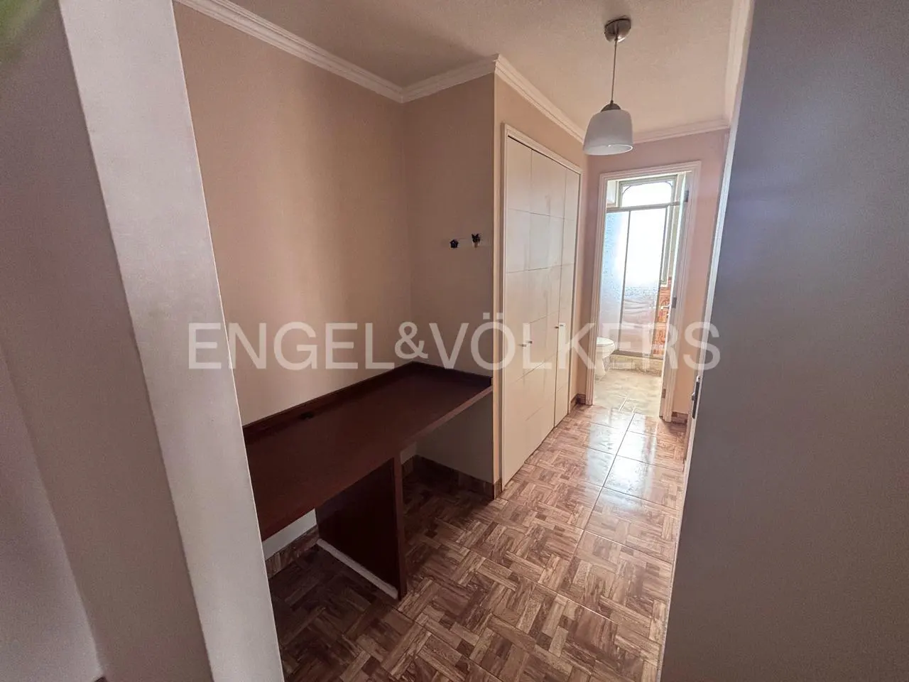 A small room with a built-in desk, closet, and patterned tile floor. A doorway leads to a bathroom. A light fixture hangs from the ceiling.