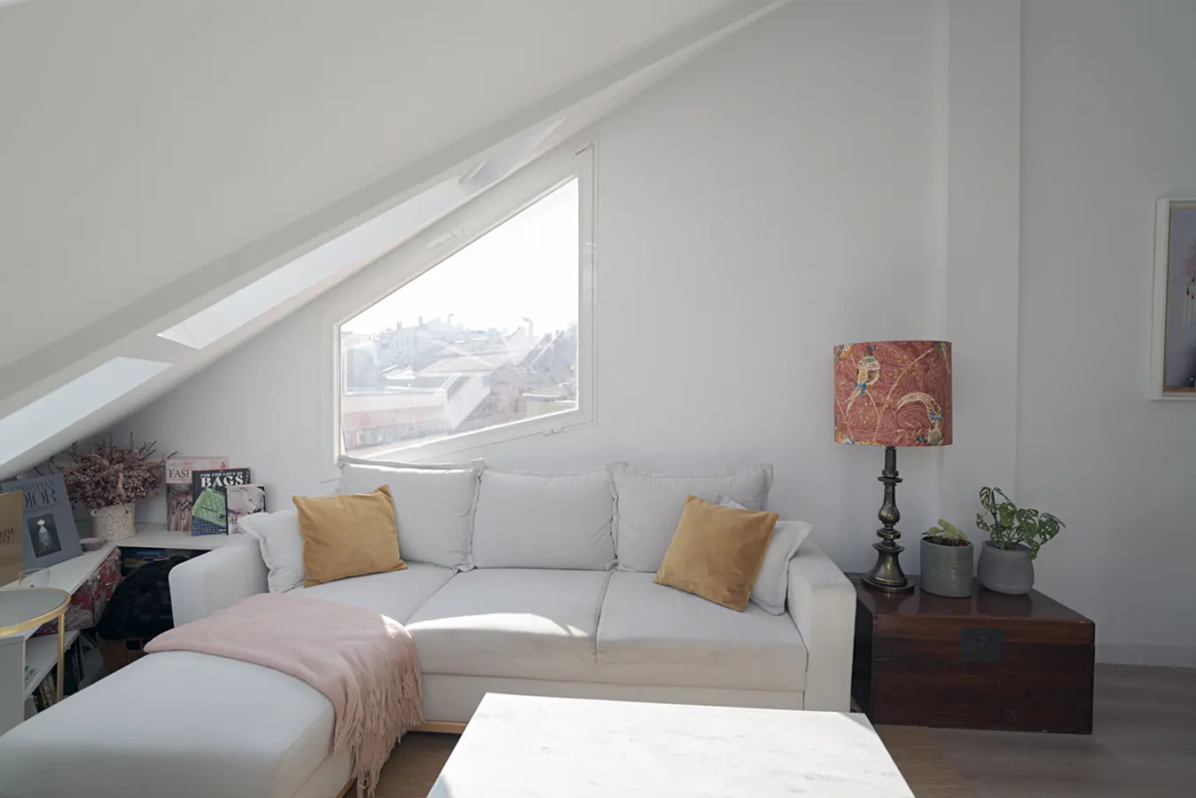 Bright, modern attic living room with white walls, a large window, and a white sectional sofa with gold pillows and a pink throw.