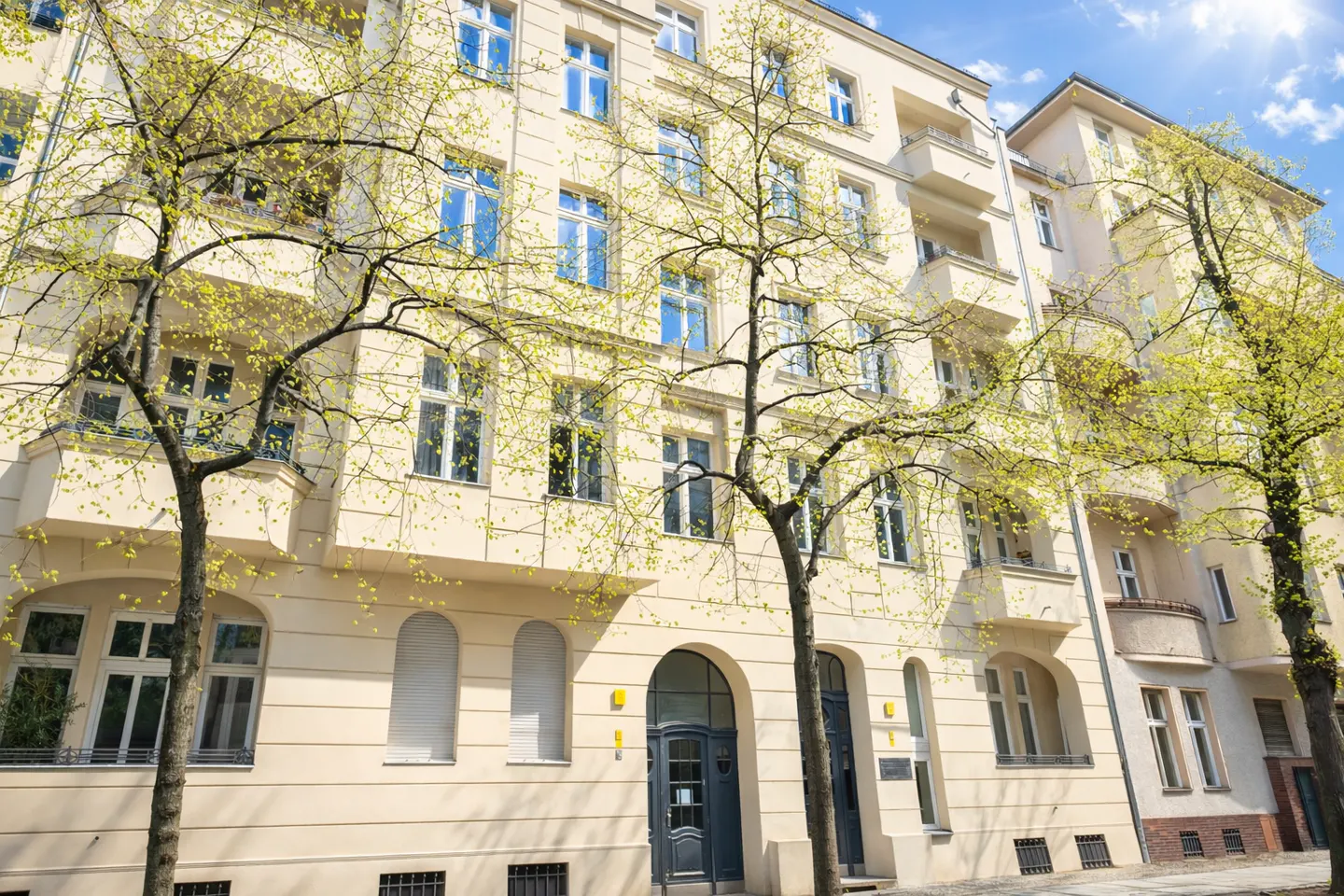 Cream-colored apartment building with many windows and balconies. Trees with green leaves frame the building. Blue sky.