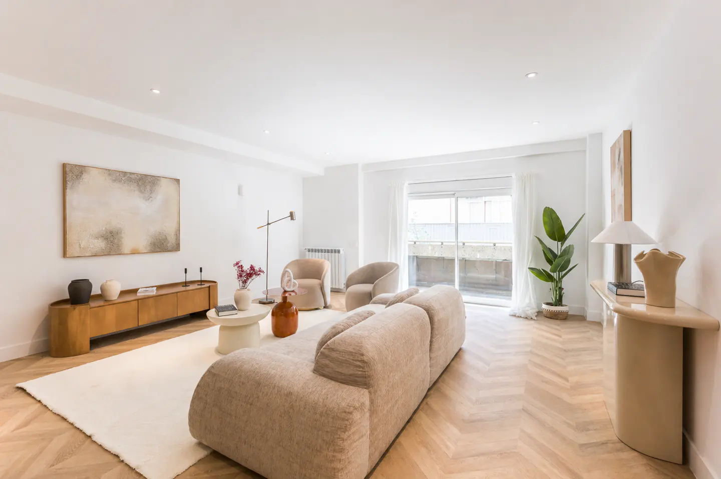 Bright living room with beige sofa, chairs, and wood floors. A large painting hangs above a wooden console. Balcony doors let in natural light.