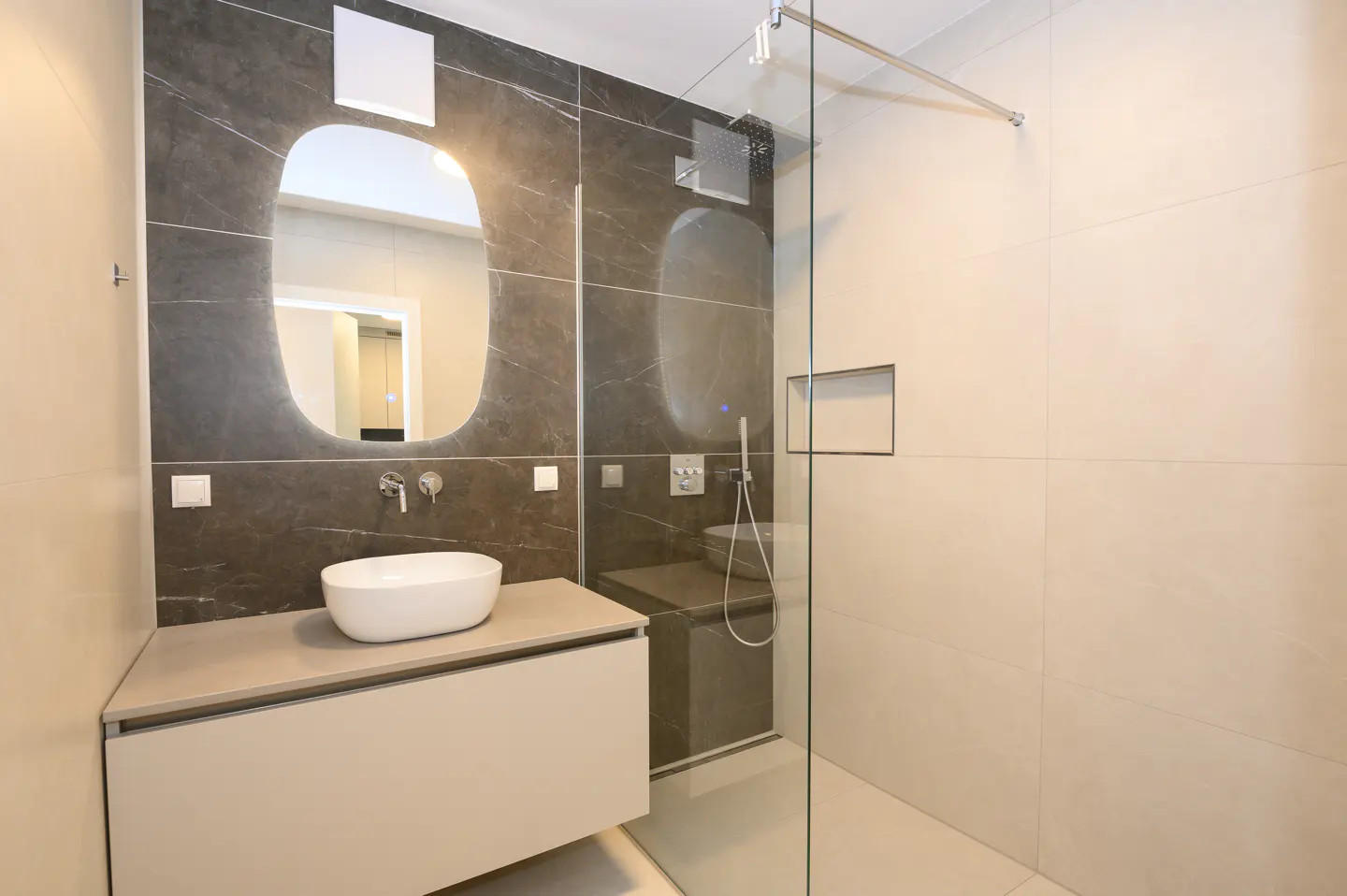 Modern bathroom with a white sink on a beige vanity, a gray tiled wall with a mirror, and a glass-enclosed shower.