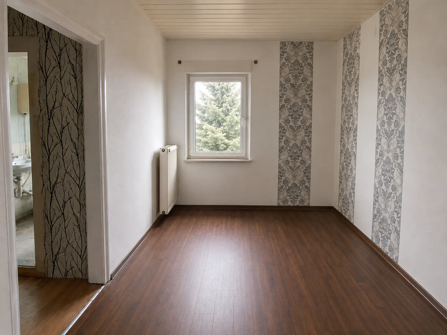 Empty room with dark wood floors, white walls, patterned wallpaper accents, a window, and a doorway to a small bathroom.