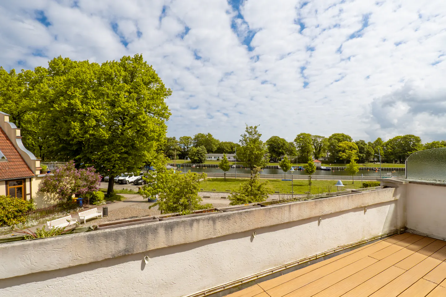 View from a balcony overlooking a green park, a calm river, and lush trees under a bright blue sky with scattered clouds.