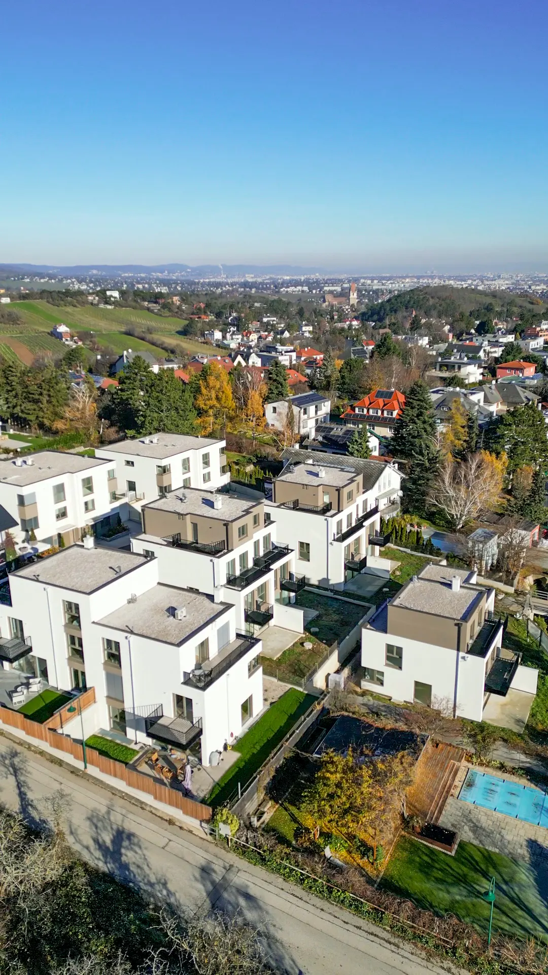 Aerial view of modern white townhouses with balconies, green lawns, and a distant cityscape under a clear blue sky.