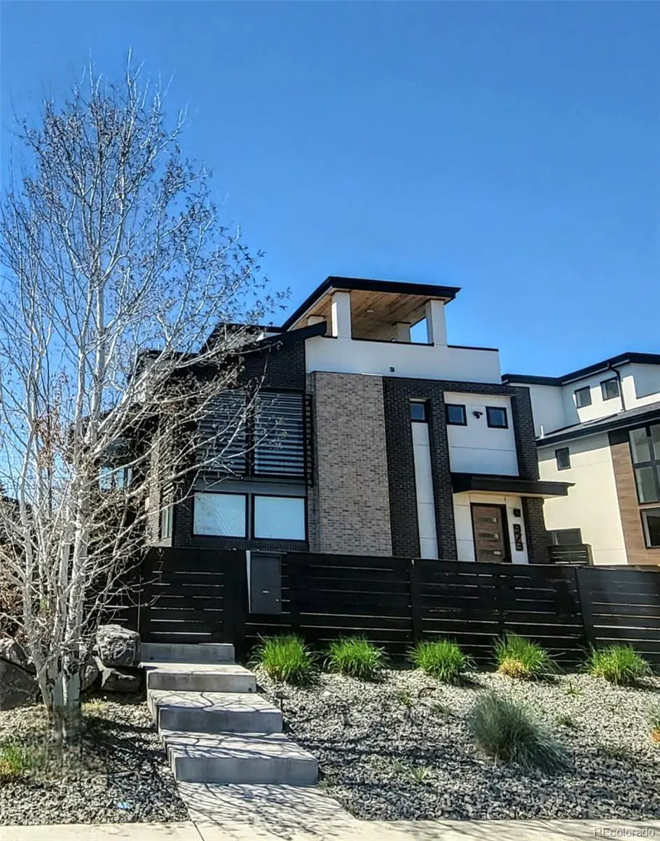 Modern two-story house with black brick, white accents, and a rooftop deck under a clear blue sky. A bare tree stands to the left.