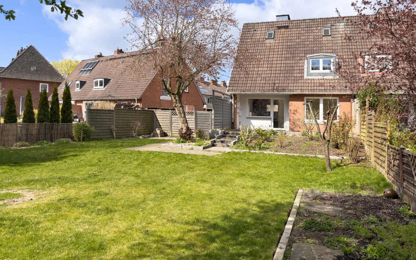 A backyard with a green lawn, a brick house with a brown roof, and a wooden fence on a sunny day.