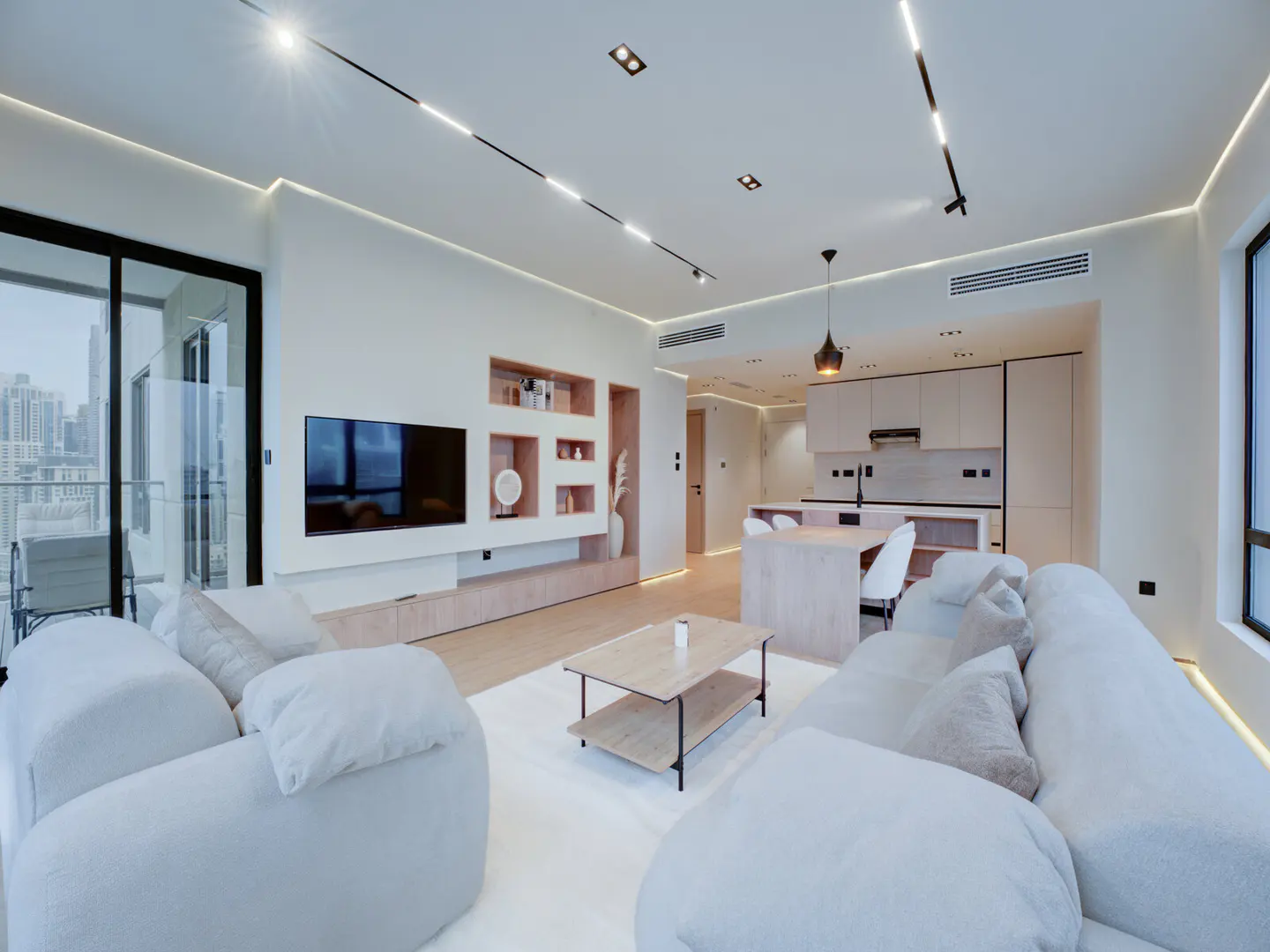 Bright, modern apartment interior with white sofas, a light wood coffee table, and a TV wall unit. The kitchen is visible in the background.