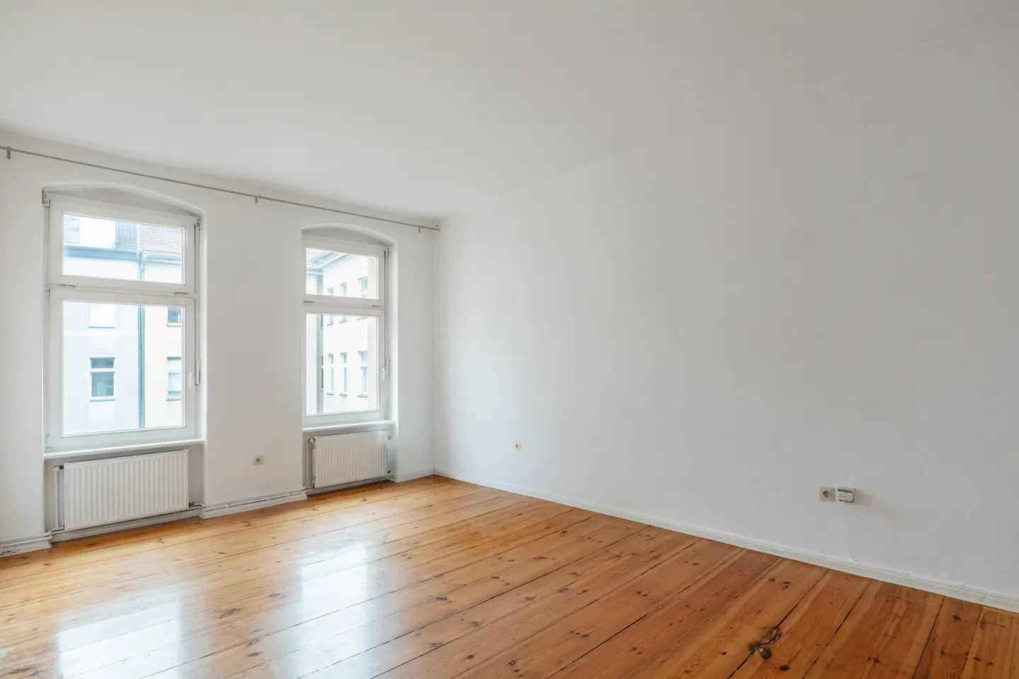 Empty room with white walls, polished wooden floors, and two large windows with white radiators underneath.
