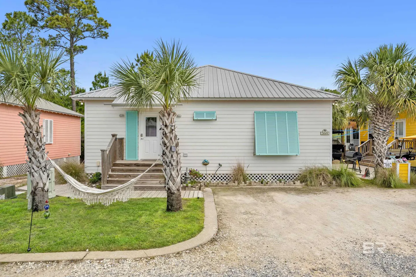 A white beach house with turquoise shutters and door, palm trees, and a hammock.