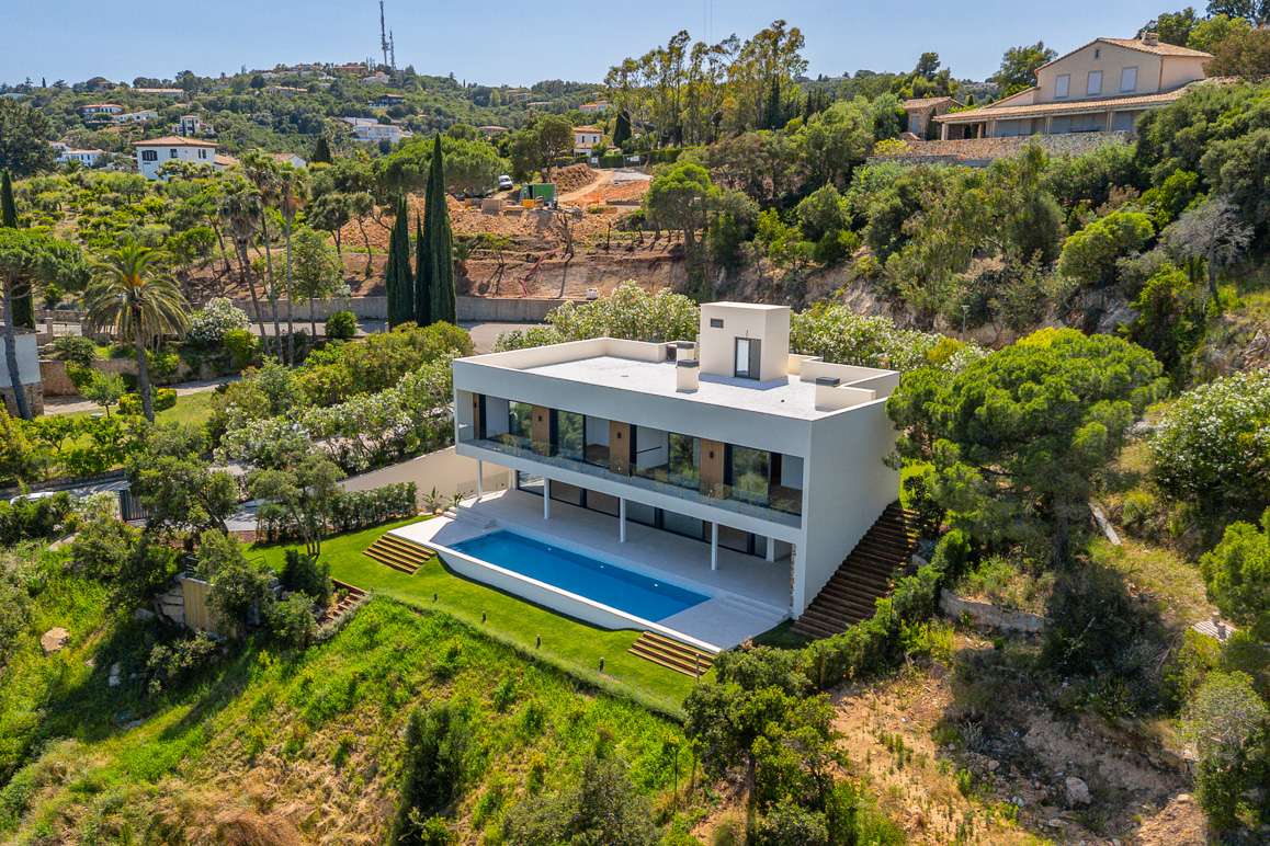 Aerial view of a modern white two-story villa with a rectangular swimming pool, set on a lush, hilly Mediterranean landscape.