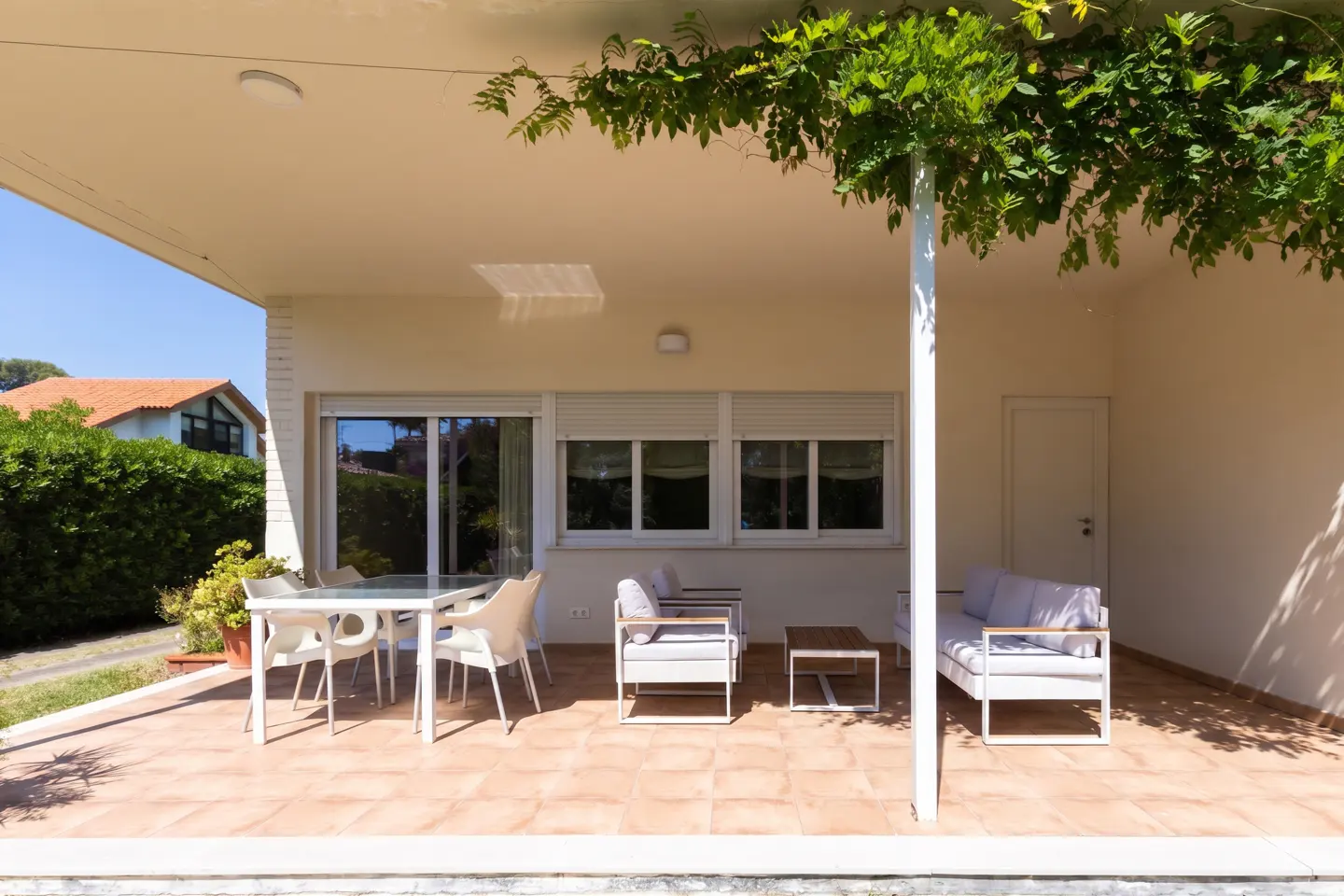 Covered patio with white furniture, including a table, chairs, and a sofa. Green vines hang from the roof. Tiled floor. Windows with closed blinds.