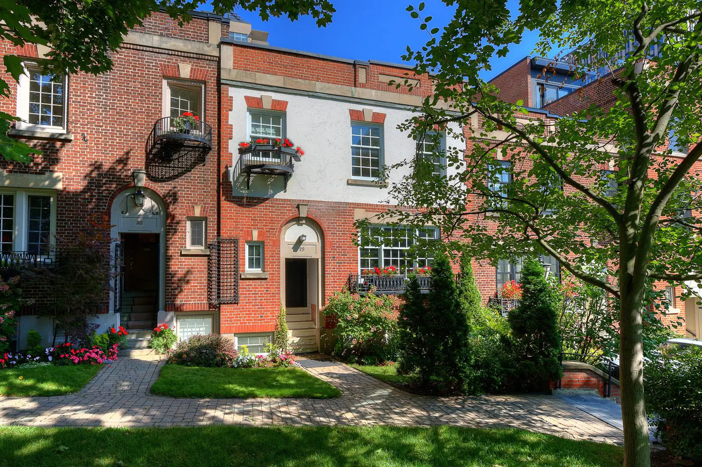 Brick townhouses with arched doorways, flower boxes, and small balconies line a green lawn under a blue sky.