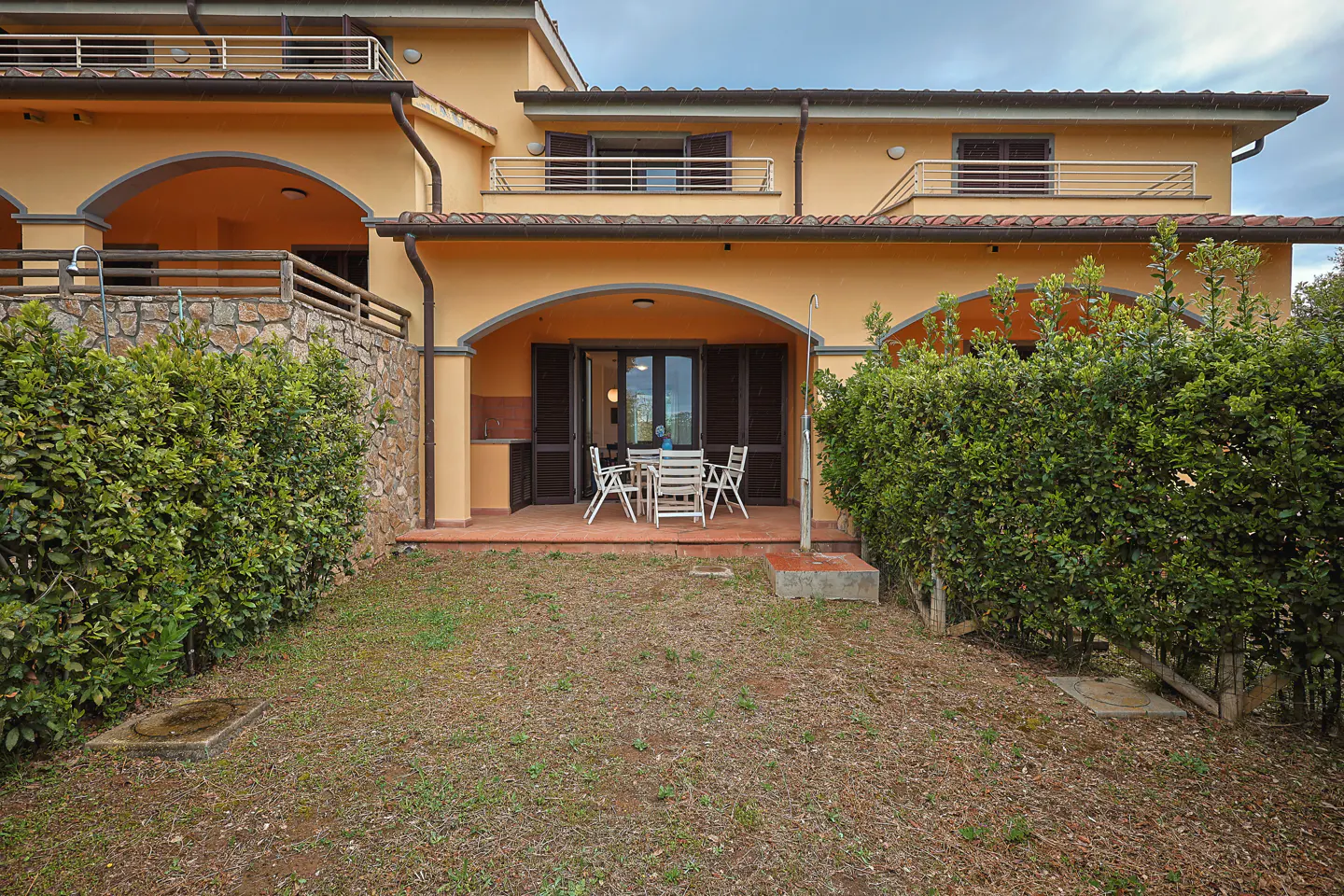 Two-story peach house with balconies and a patio with a white table and chairs. Green bushes surround the patio.